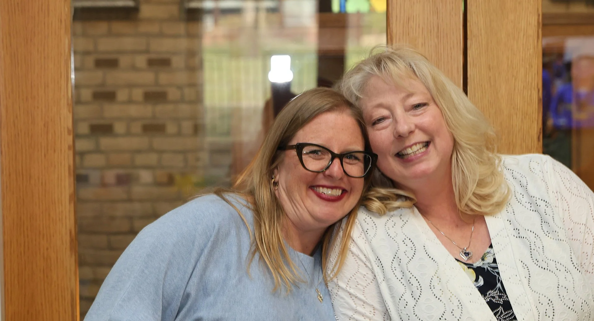 Two women smiling and hugging each other, standing near a wooden-framed glass partition with a brick wall in the background.