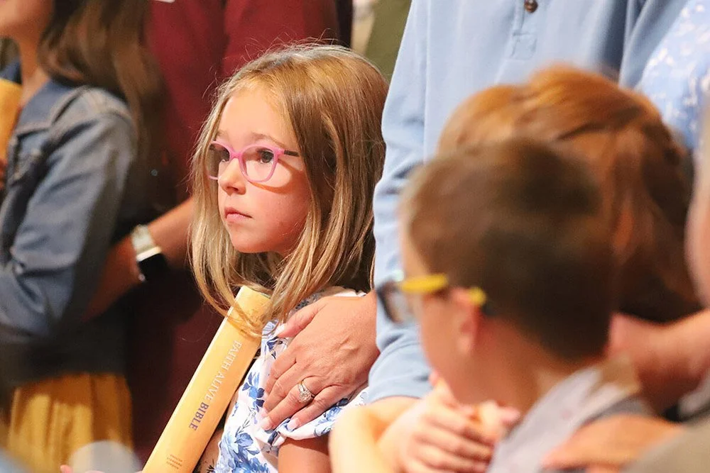 A young girl with red hair and glasses holding a yellow Bible at a church service surrounded by other children and adults.