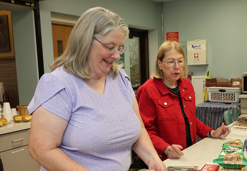 Two middle-aged women standing at a food counter, smiling and looking down, with various food items and snack packages on the counter.