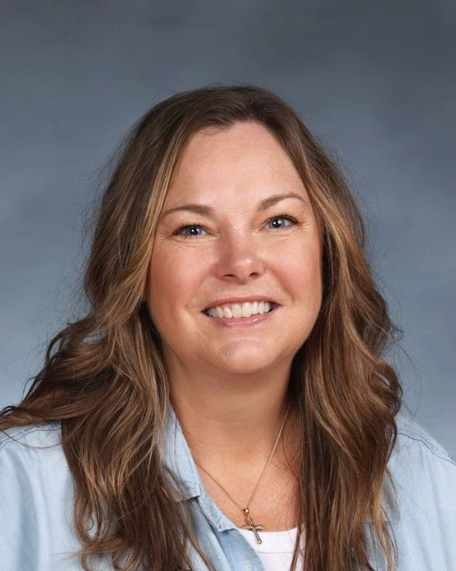 Smiling woman with long, wavy brown hair wearing a light blue shirt and a gold cross necklace, against a gray background.
