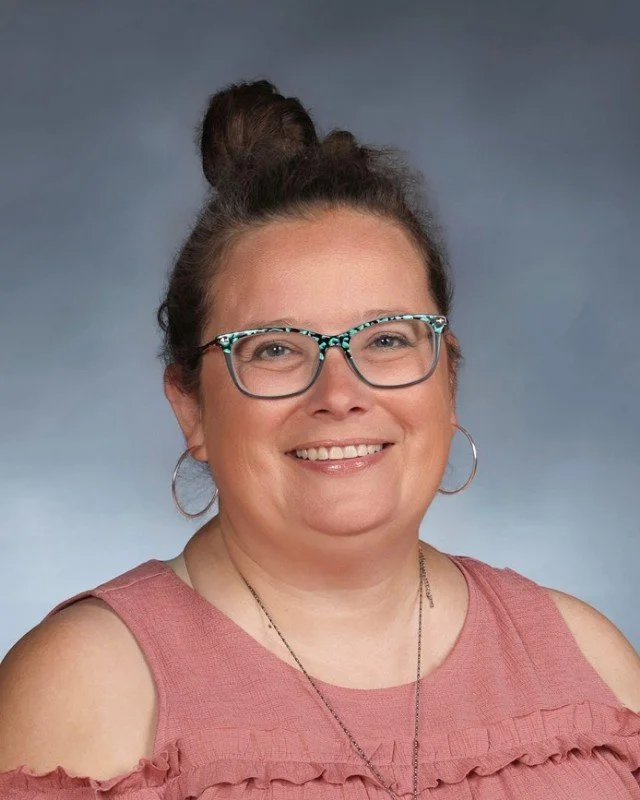 A woman with glasses, hoop earrings, and a pink sleeveless top, smiling against a bluish-gray background.
