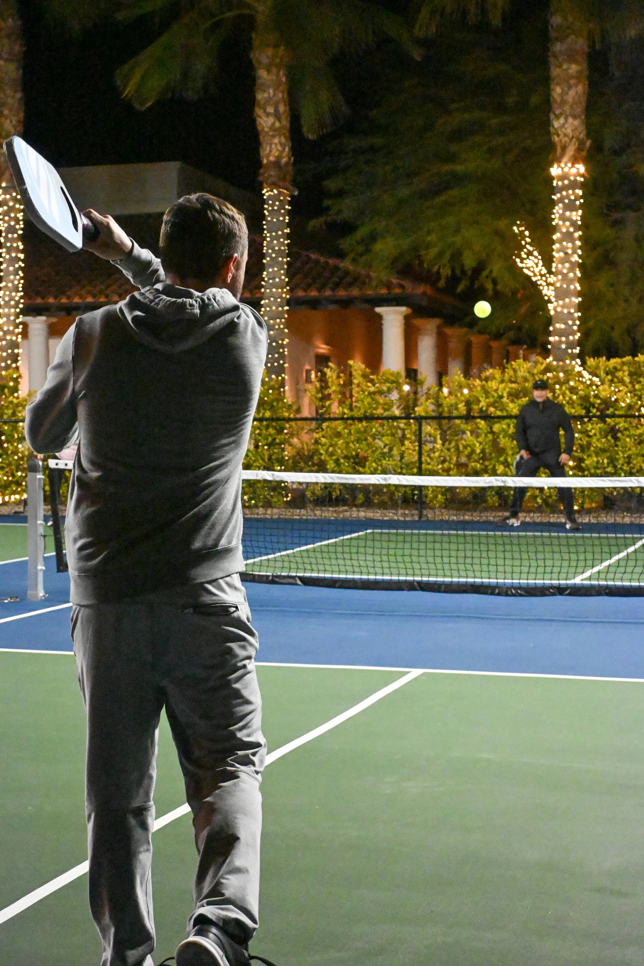 Two people playing pickleball on an outdoor court at night, with trees and decorative lights in the background.