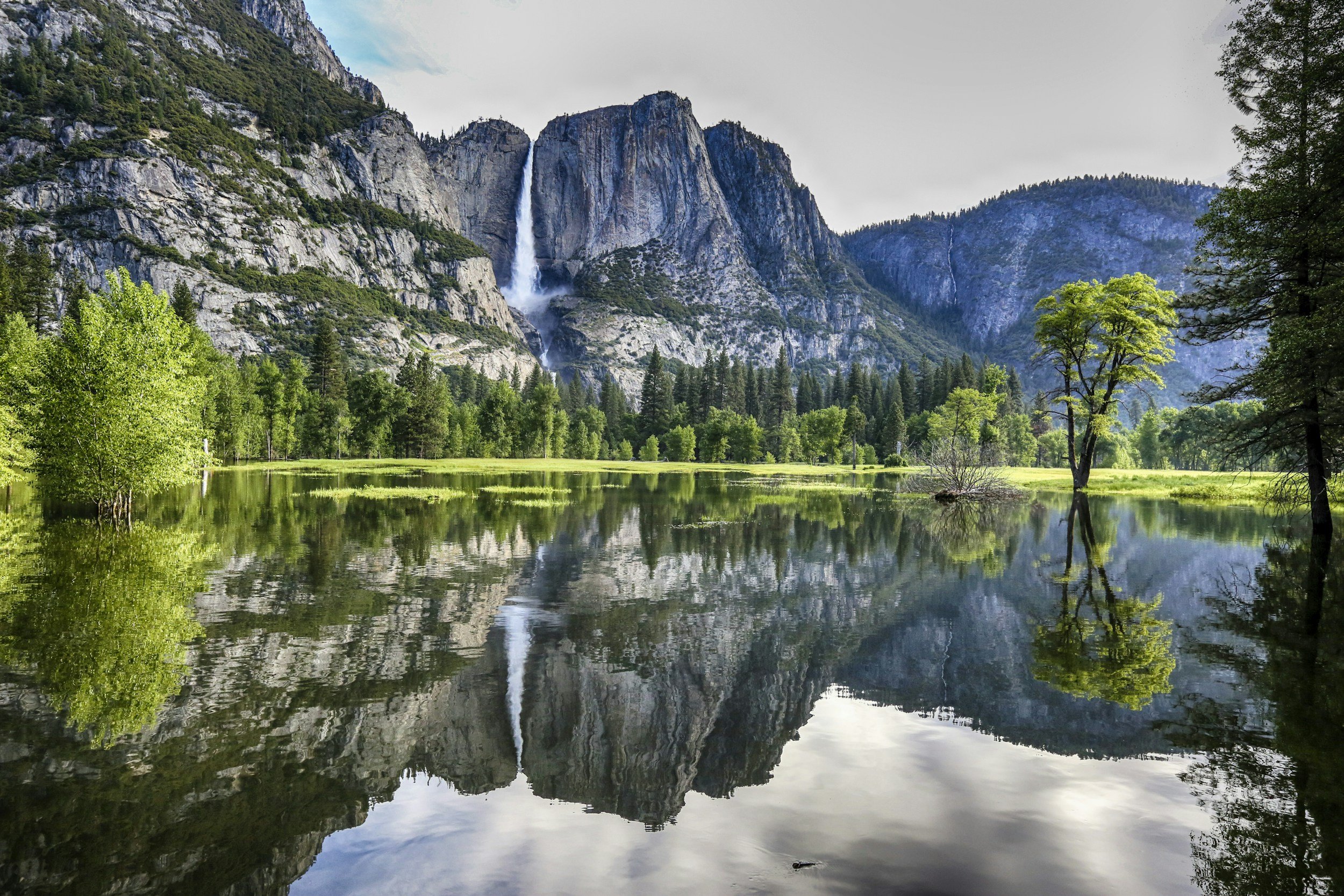 A serene landscape of a mountain lake with lush green trees, reflecting the surrounding mountains and waterfalls on a clear day.