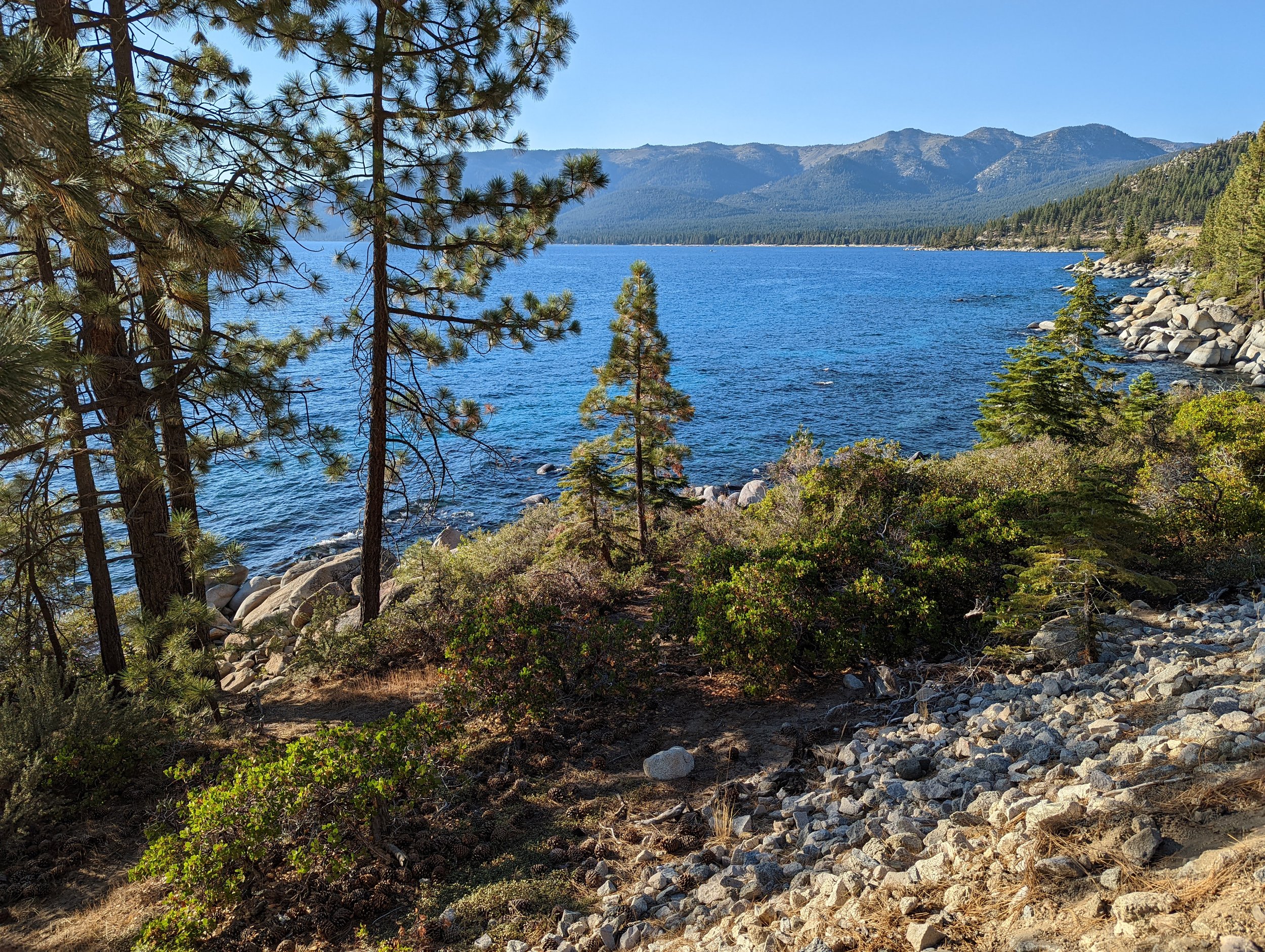 A scenic view of a lake lined with trees and rocks, with mountains in the background under a clear blue sky.