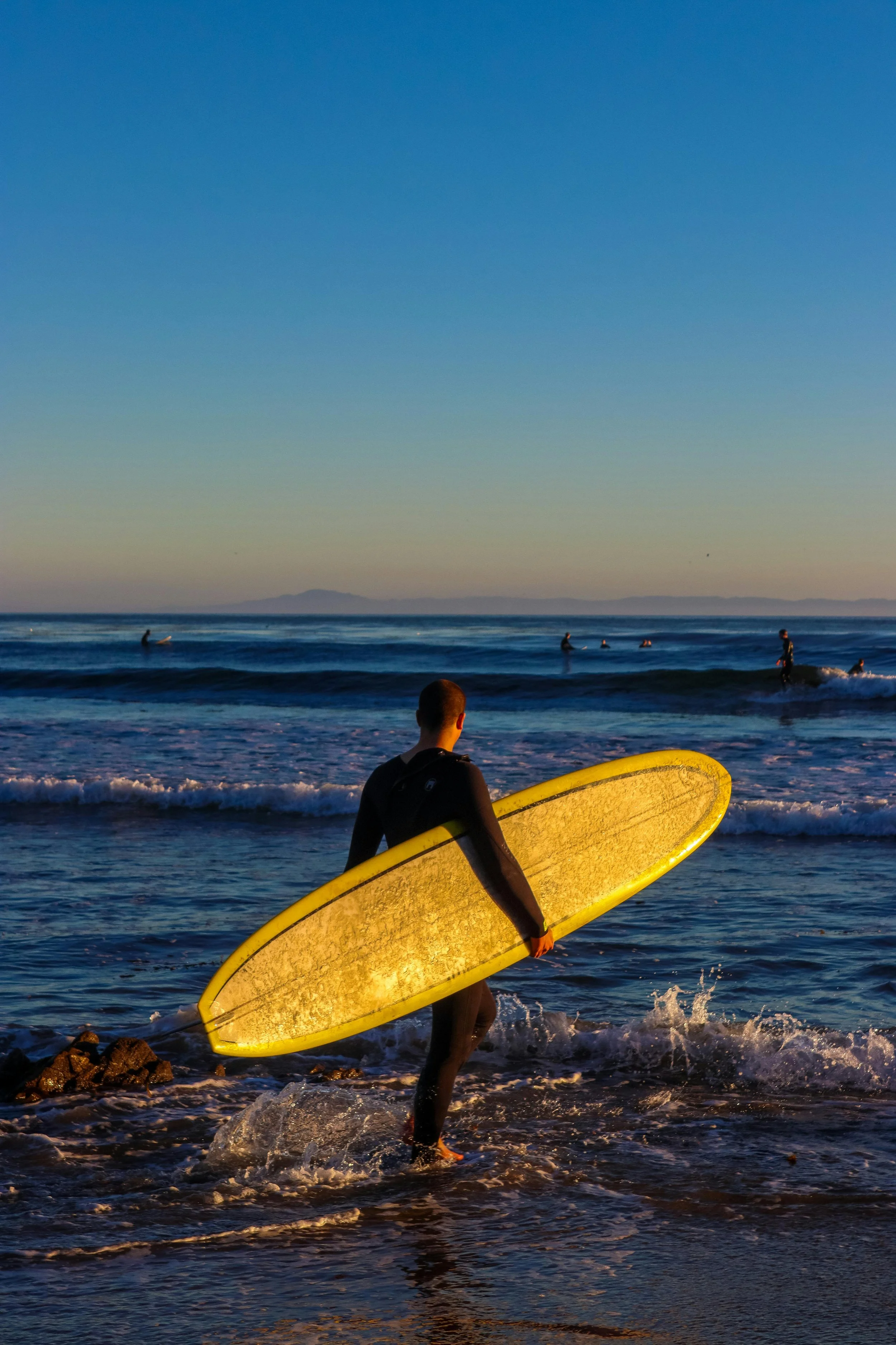 A person in a wetsuit carrying a yellow surfboard walking out of the ocean at sunset, with several people surfing in the background.