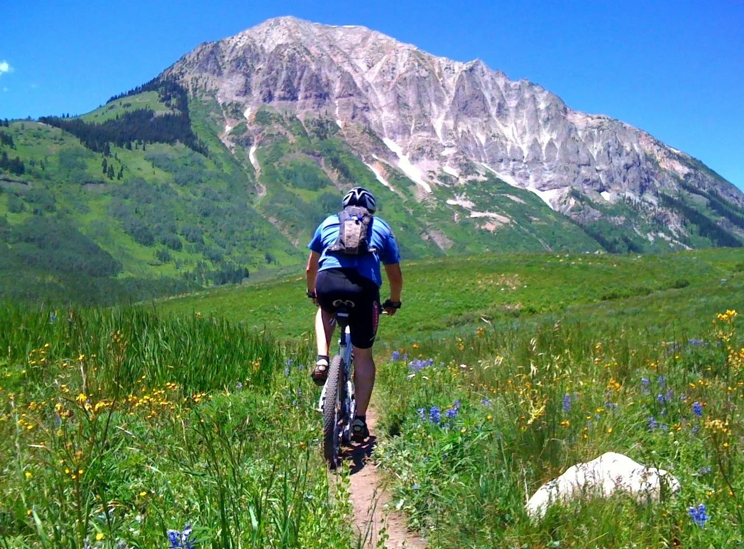A person standing with a mountain bike on a trail, overlooking a scenic mountain landscape with trees, a lake, and a partly cloudy sky during sunset.
