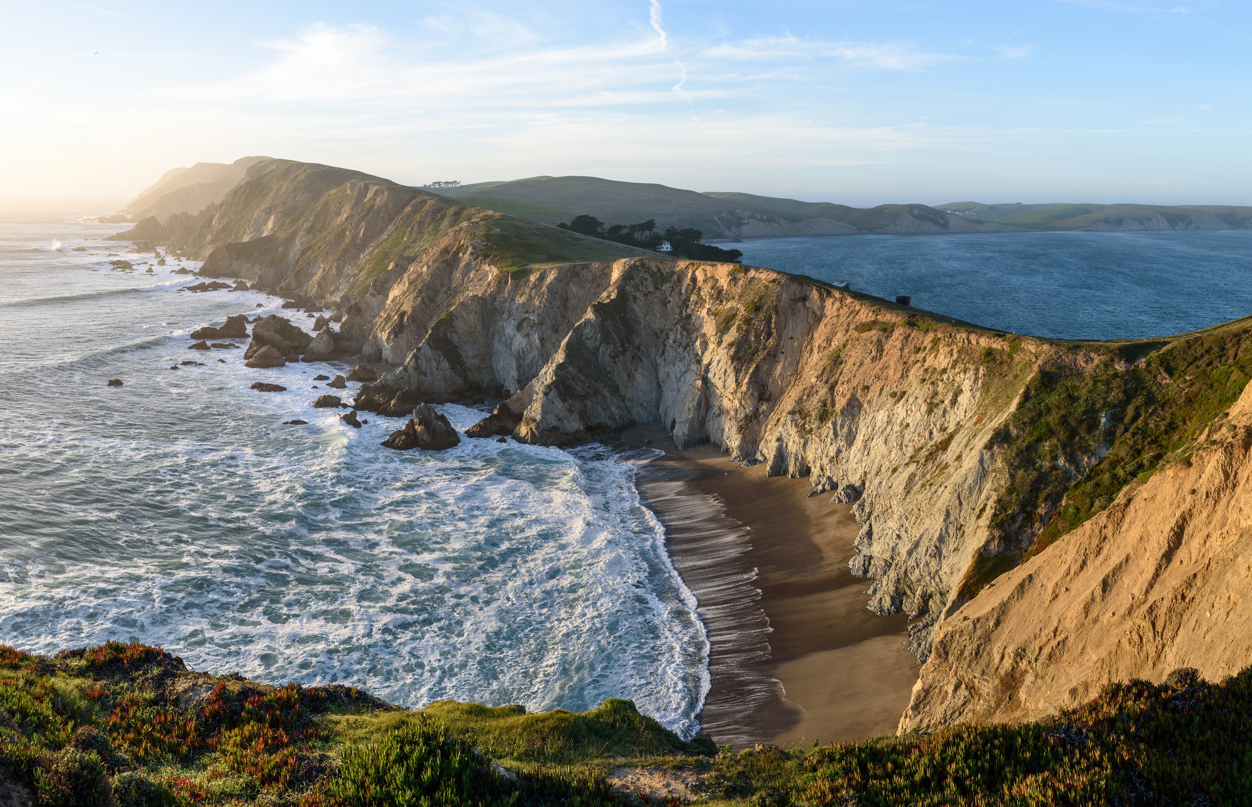 Coastal cliffs and beach at sunset with waves crashing on shore, grassy and rocky terrain in foreground, blue ocean and sky in background.