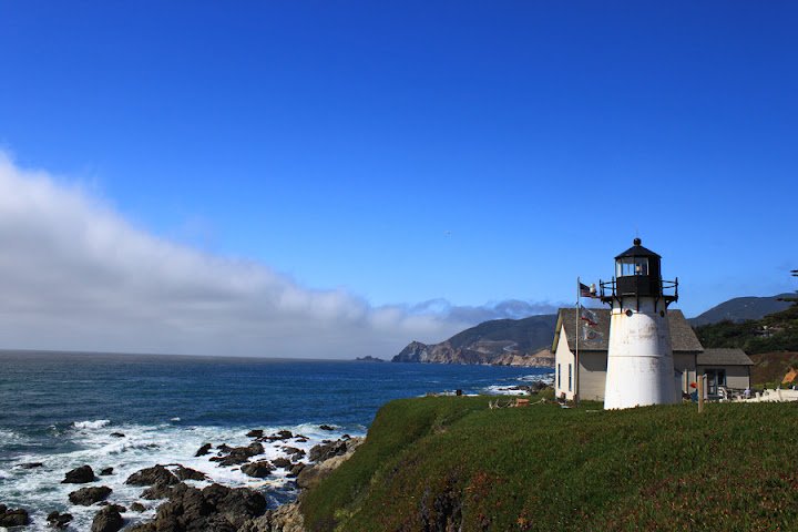 A lighthouse near the coastline with a mountain in the background, under a clear blue sky.