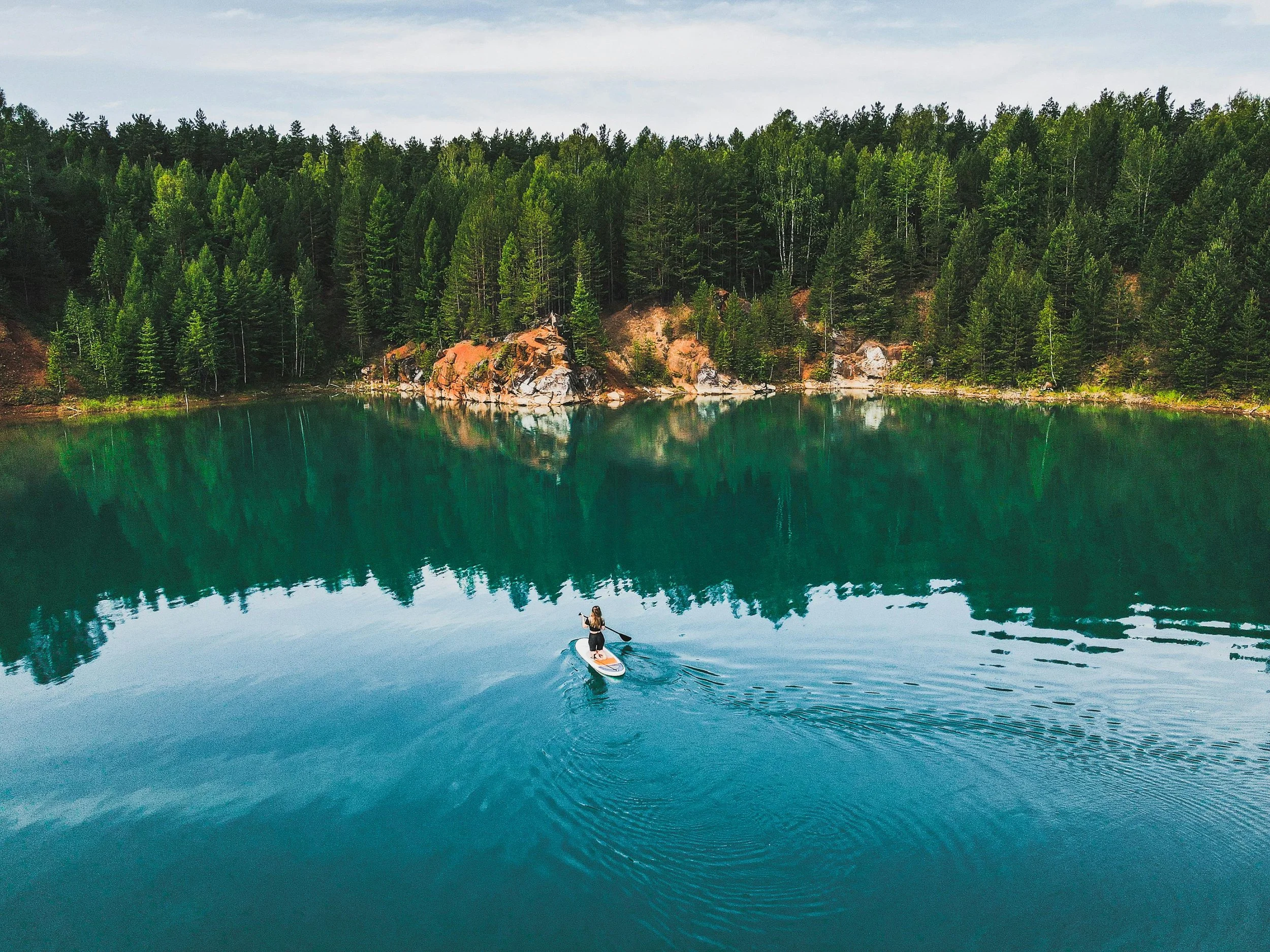 A person paddleboarding on a calm, greenish-blue lake surrounded by a dense forest of green trees.