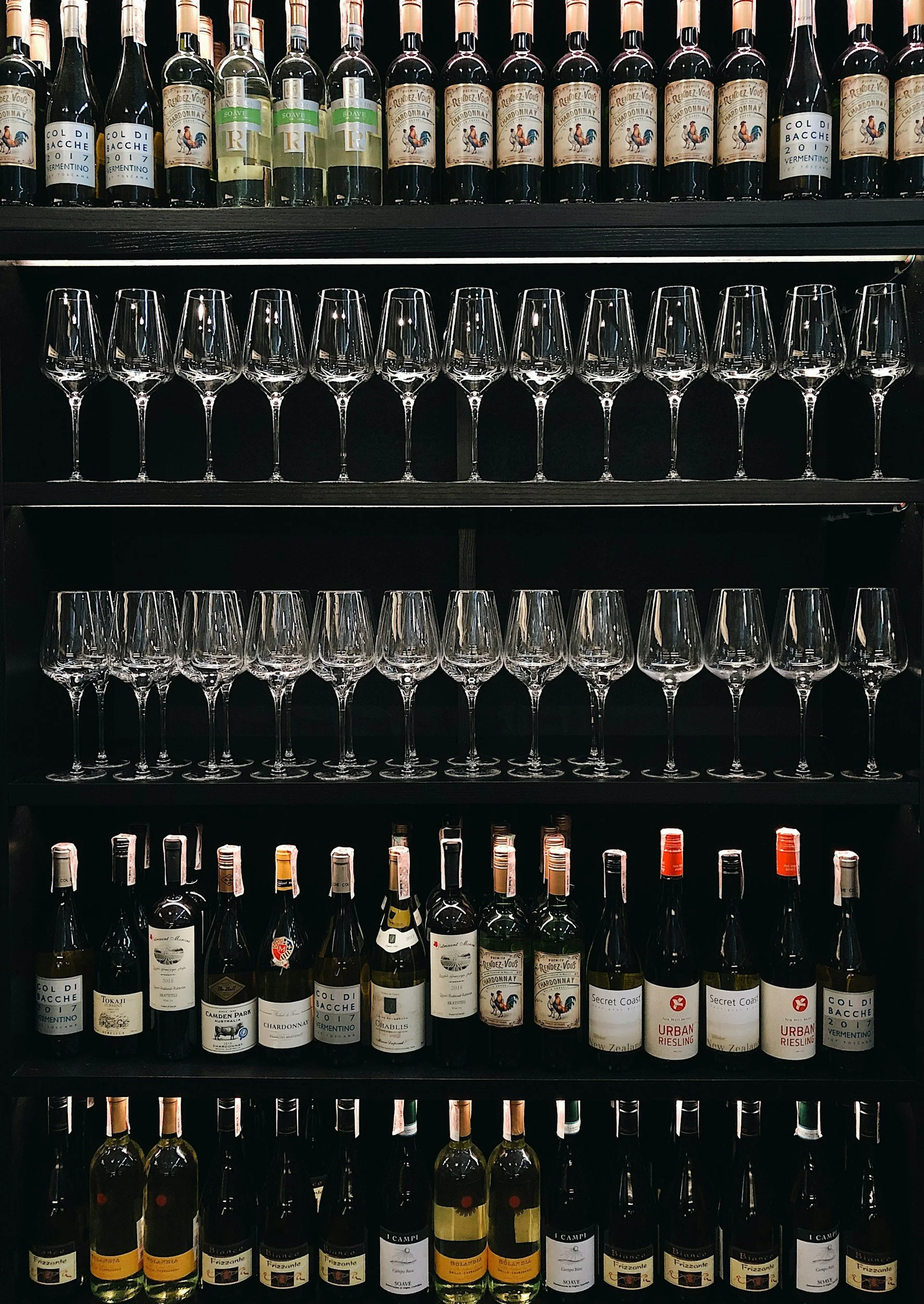 Shelf of wine bottles and glassware, including empty wine glasses and wine bottles of various labels and types arranged on black shelves.