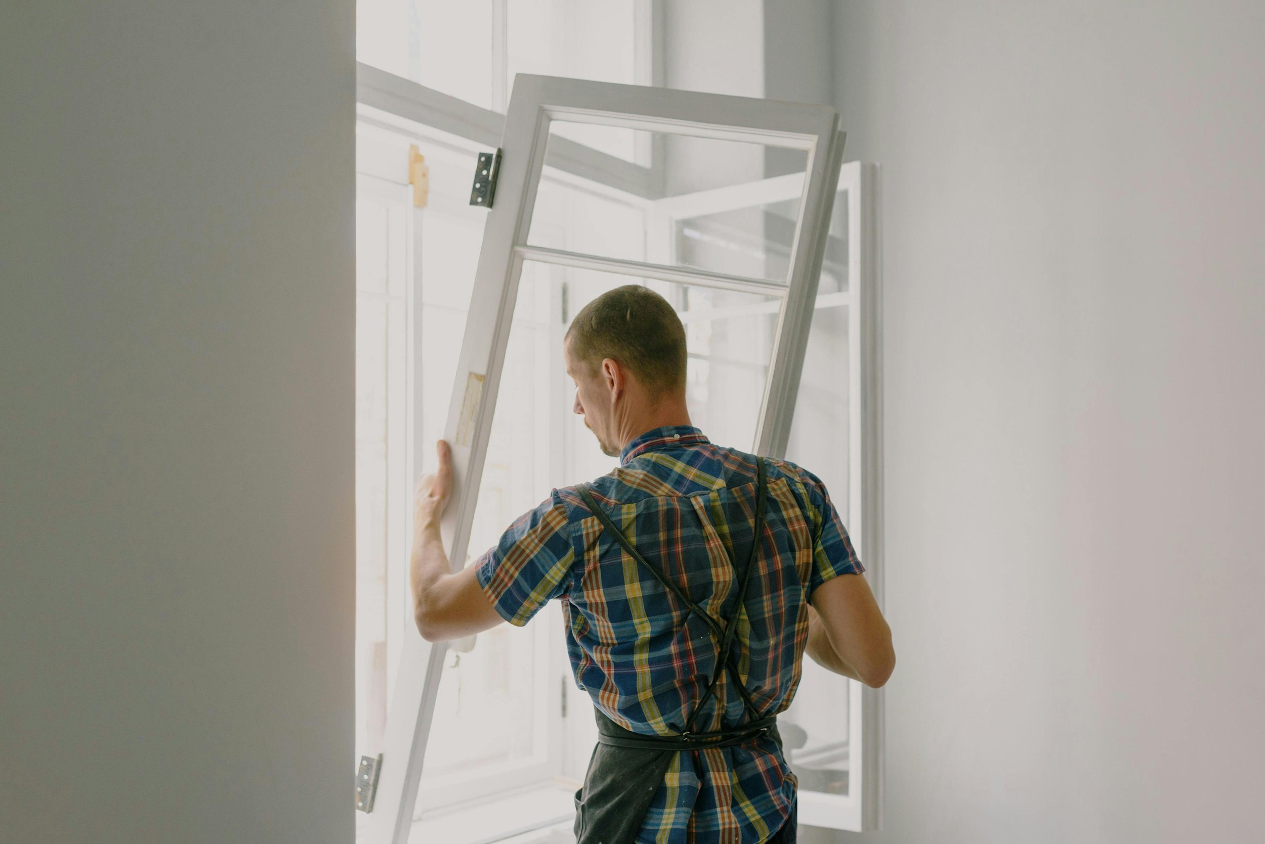 A man installing a white window frame in a room with white walls.