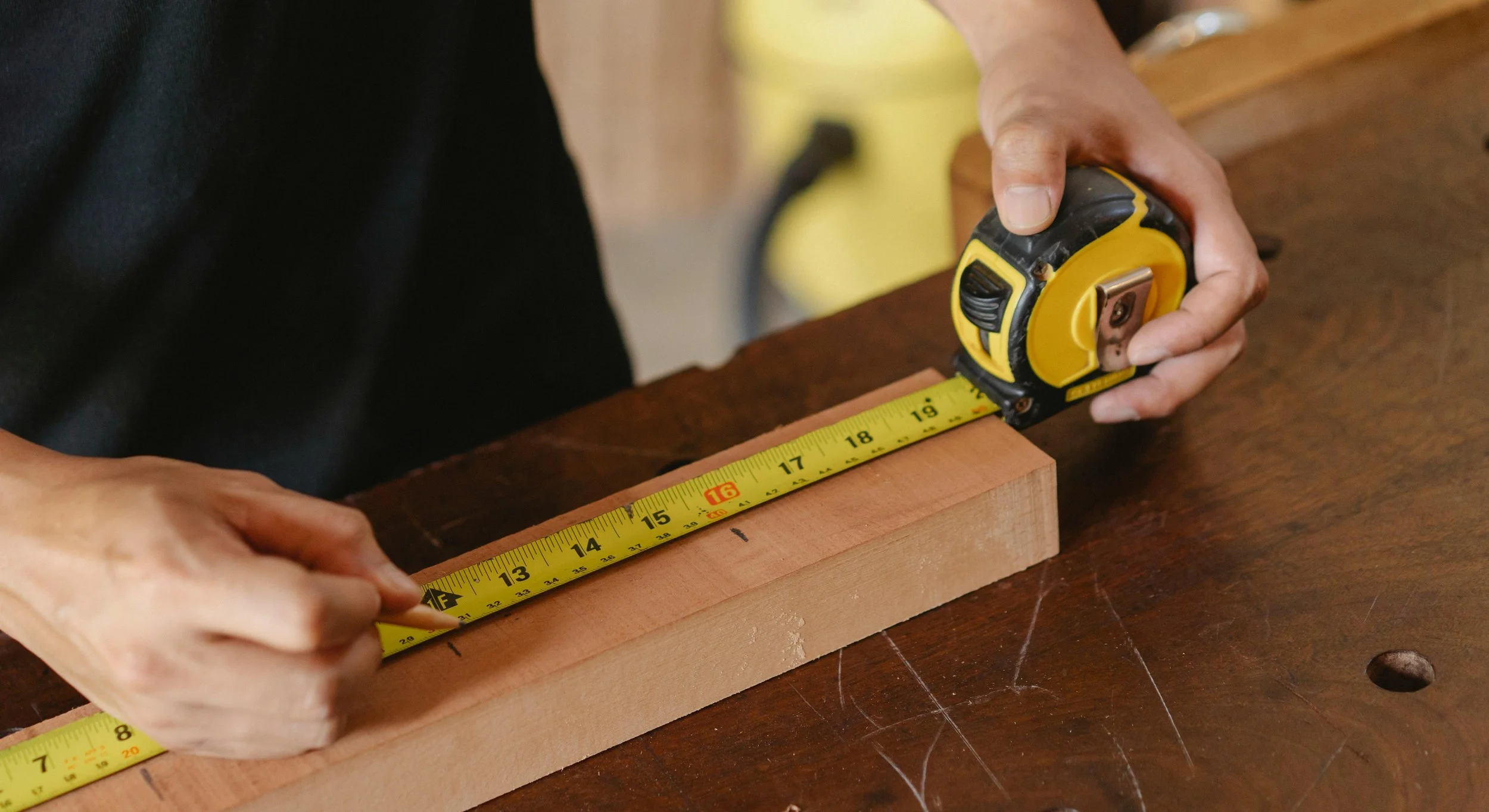 A person measuring a piece of wood on a wooden workbench with a yellow and black tape measure.