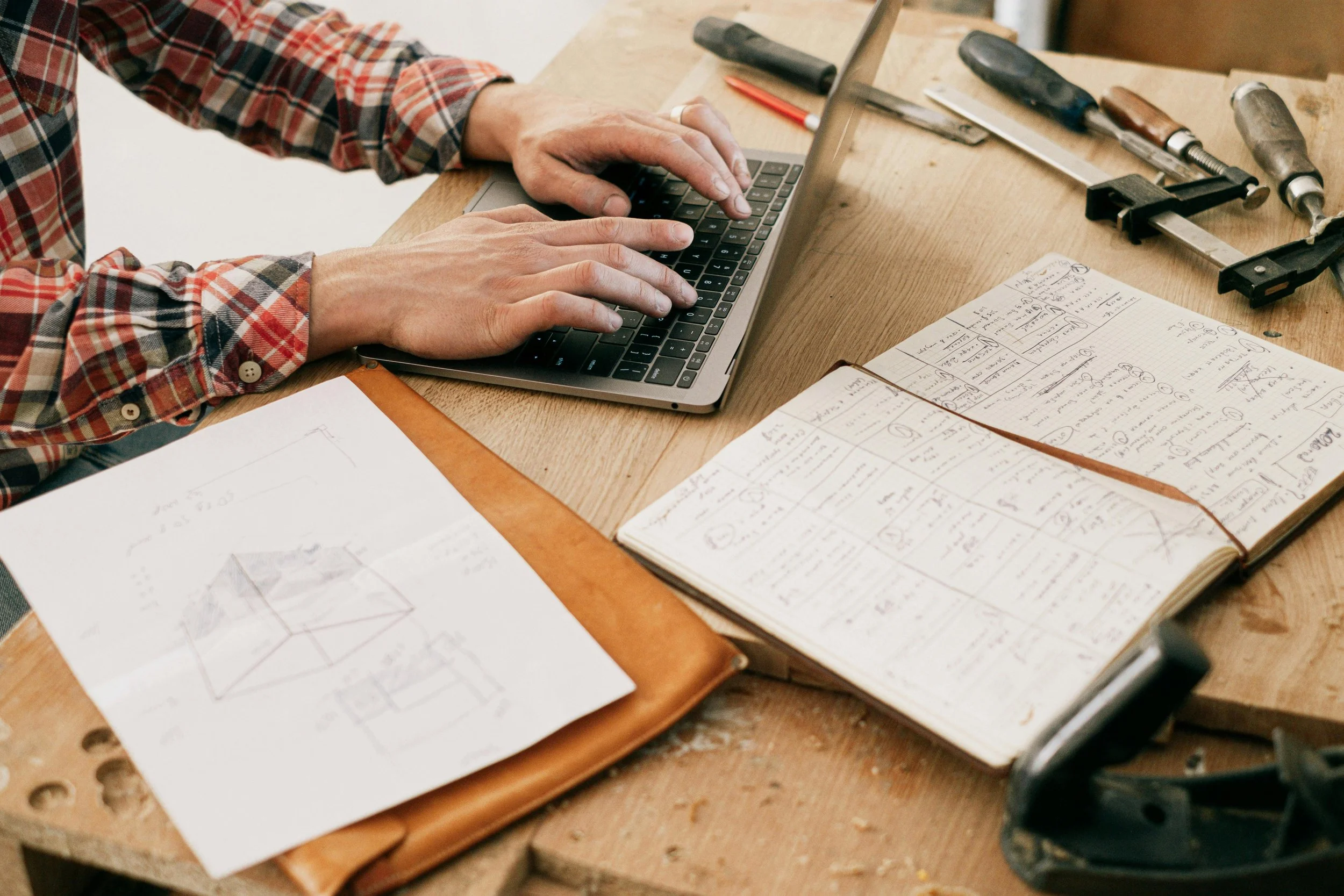 Person in a plaid shirt working on a laptop at a cluttered wooden workbench with sketches, a notebook filled with handwritten notes, and various woodworking tools.