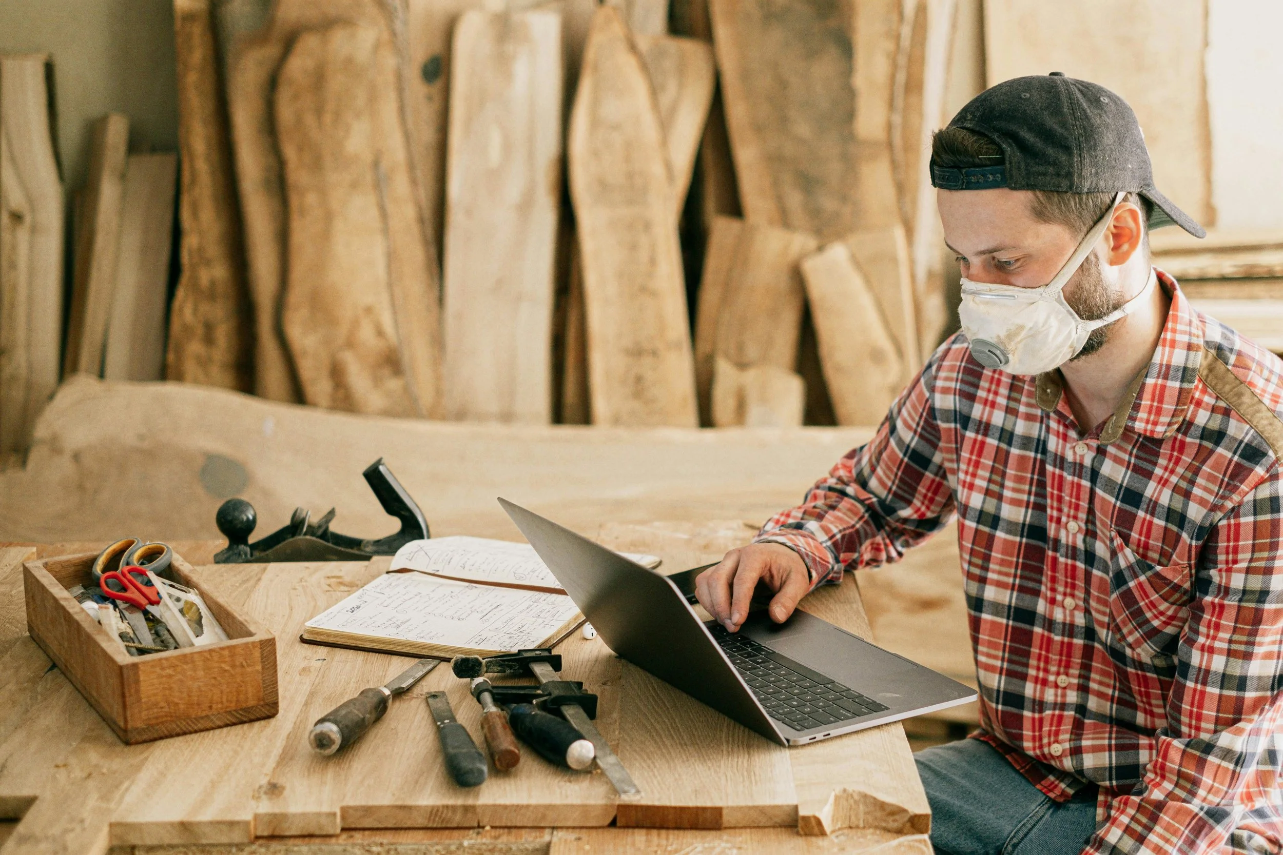 A man wearing a mask, baseball cap, and plaid shirt working on a laptop at a woodworking shop with wood planks and tools on a workbench.