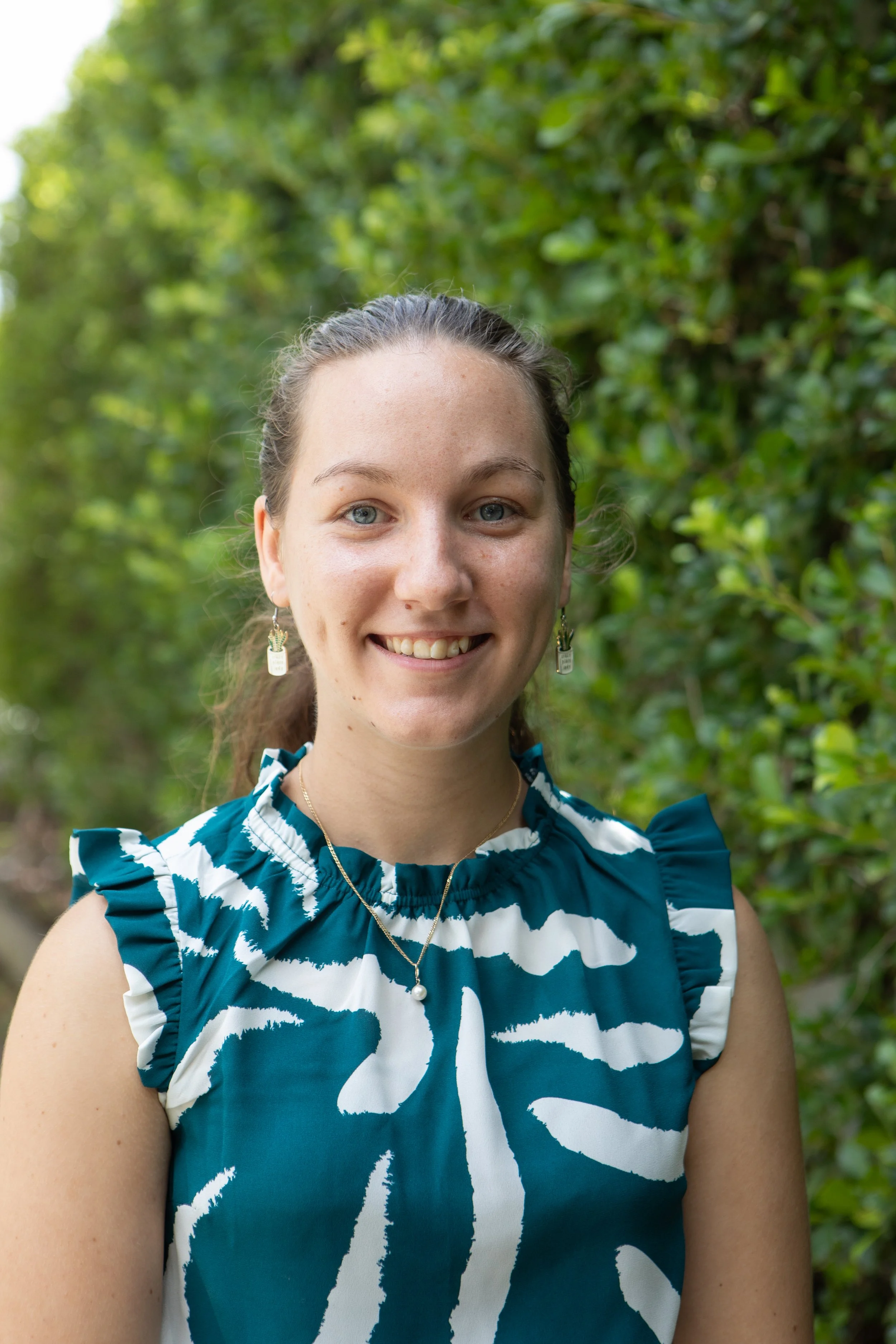 A young woman with light brown hair, wearing a blue and white patterned sleeveless top, and jewelry including earrings and a necklace, smiling outdoors in front of green leafy background.