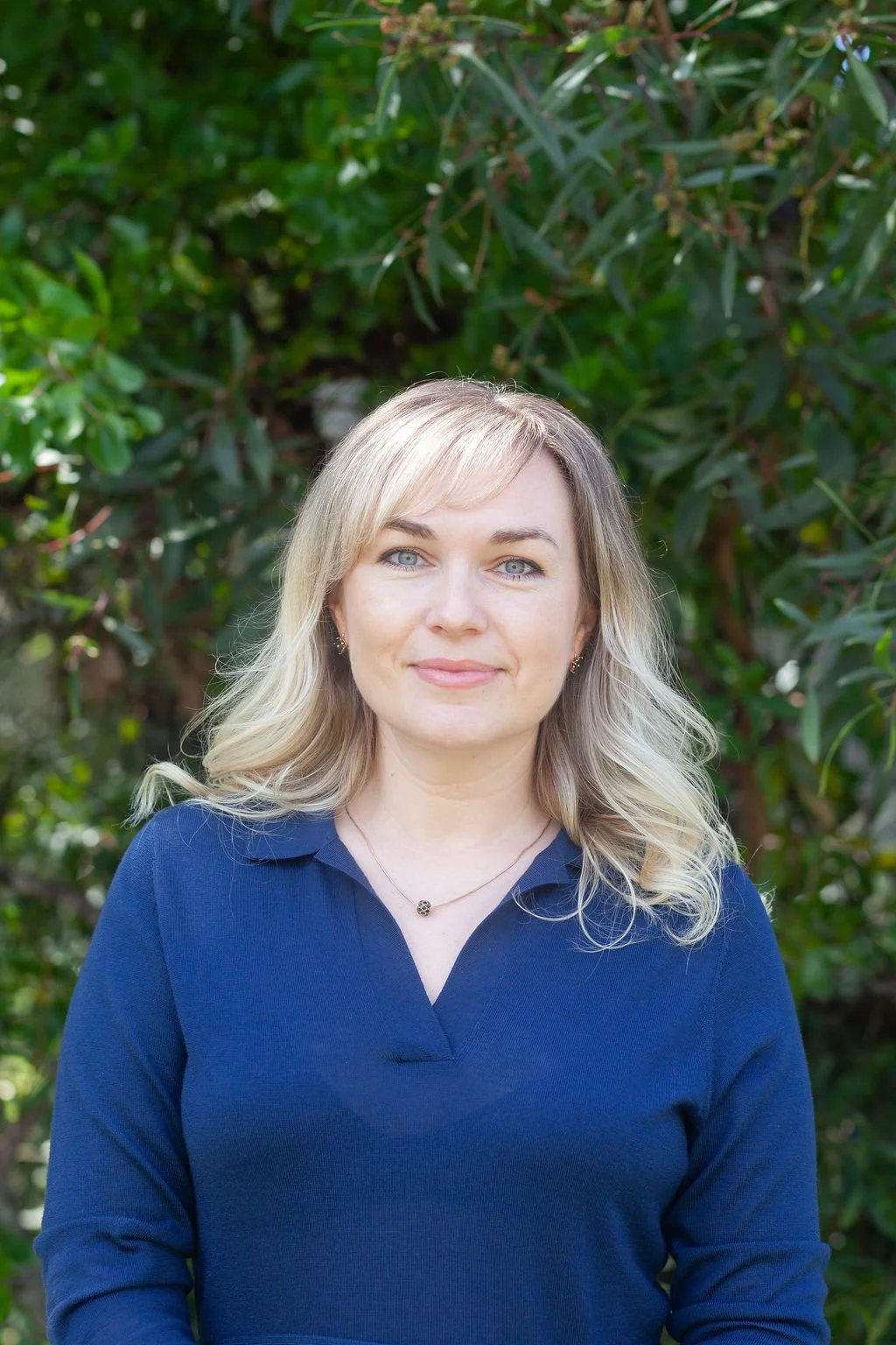A woman with blonde hair and blue eyes wearing a navy blue blouse standing outdoors in front of green foliage.