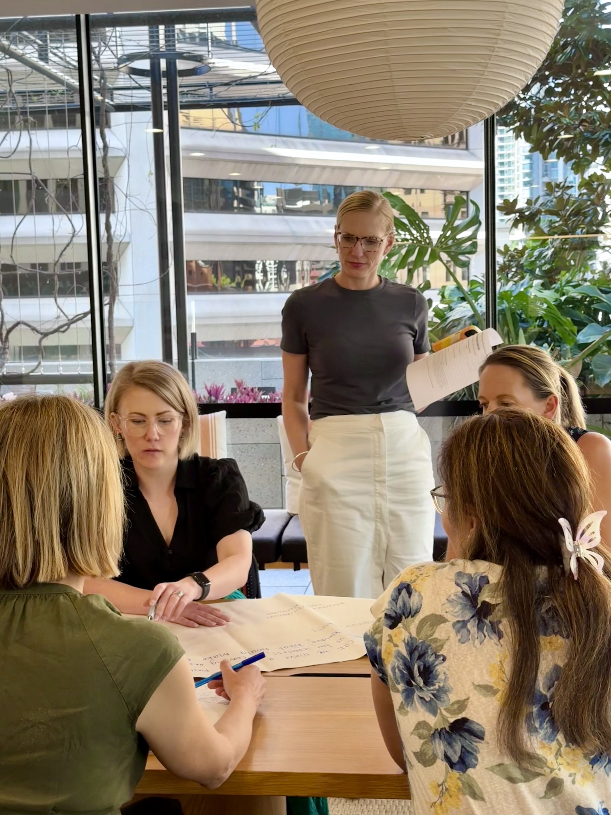 A group of women seated around a table engaged in a discussion with a woman standing, holding a notebook, in a bright room with large windows, green plants, and modern city buildings outside.