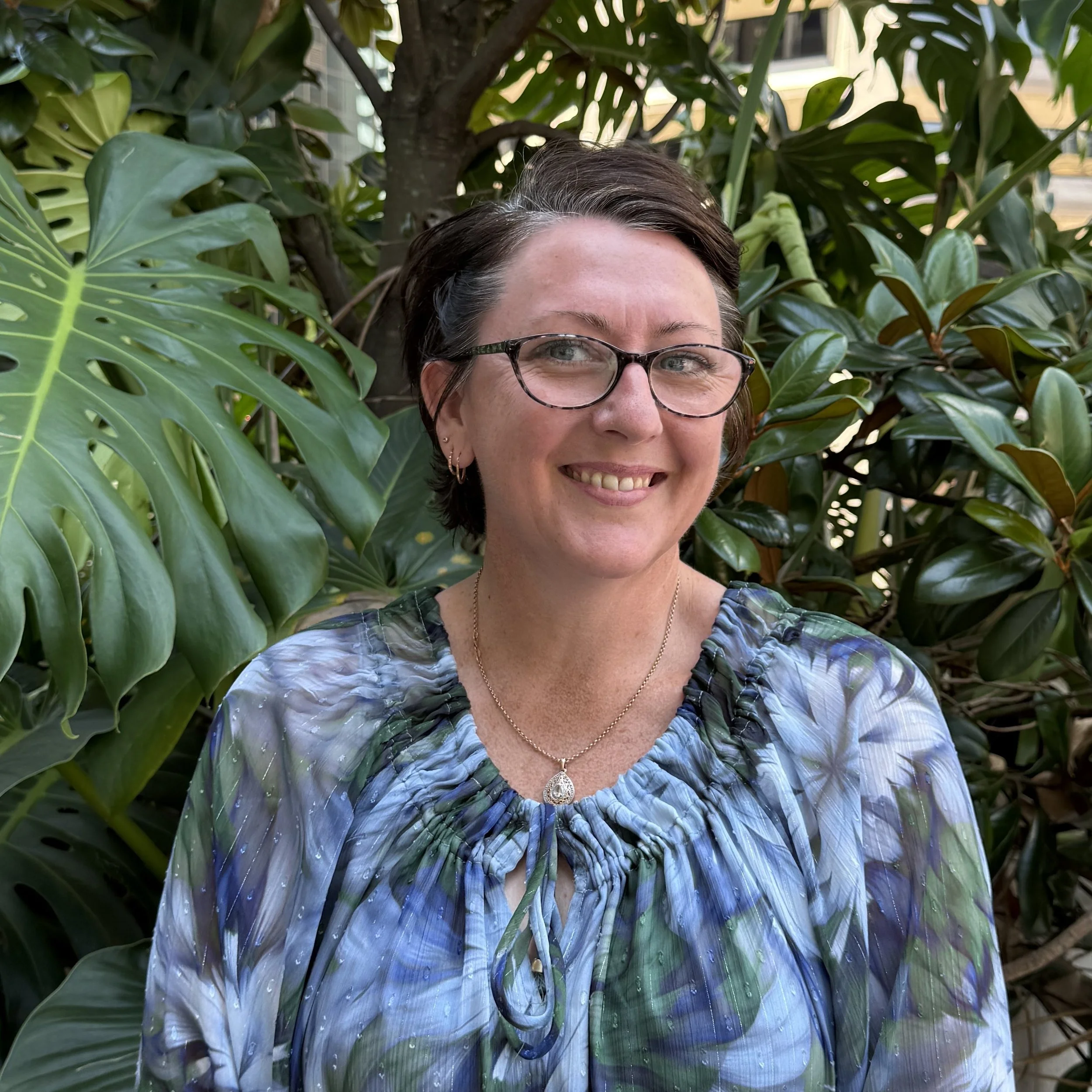 A woman with short dark hair, glasses, and a necklace, smiling and standing in front of green foliage in an outdoor setting.