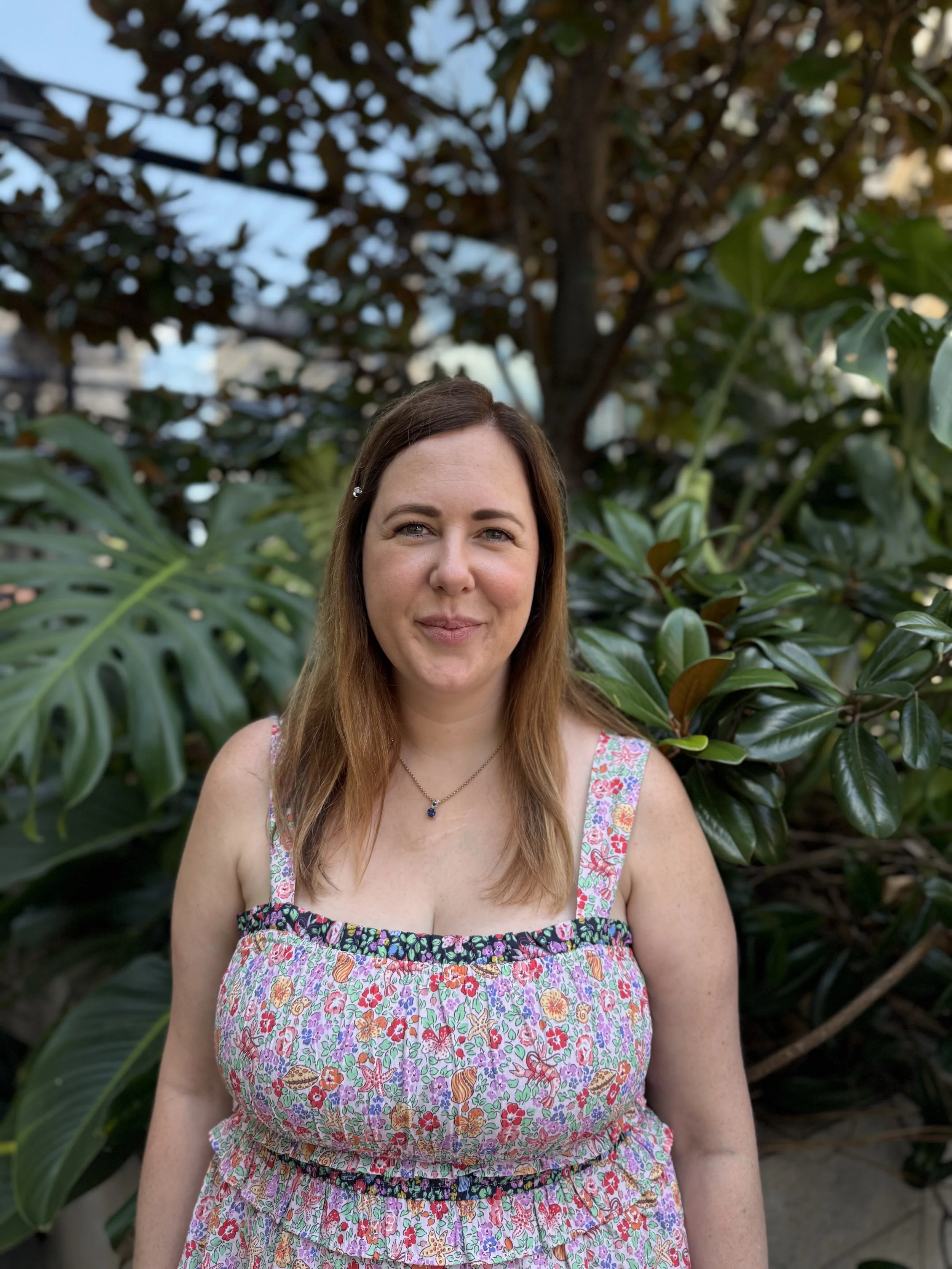 A woman with light brown hair standing outdoors in front of green tropical plants, wearing a floral dress and a necklace.