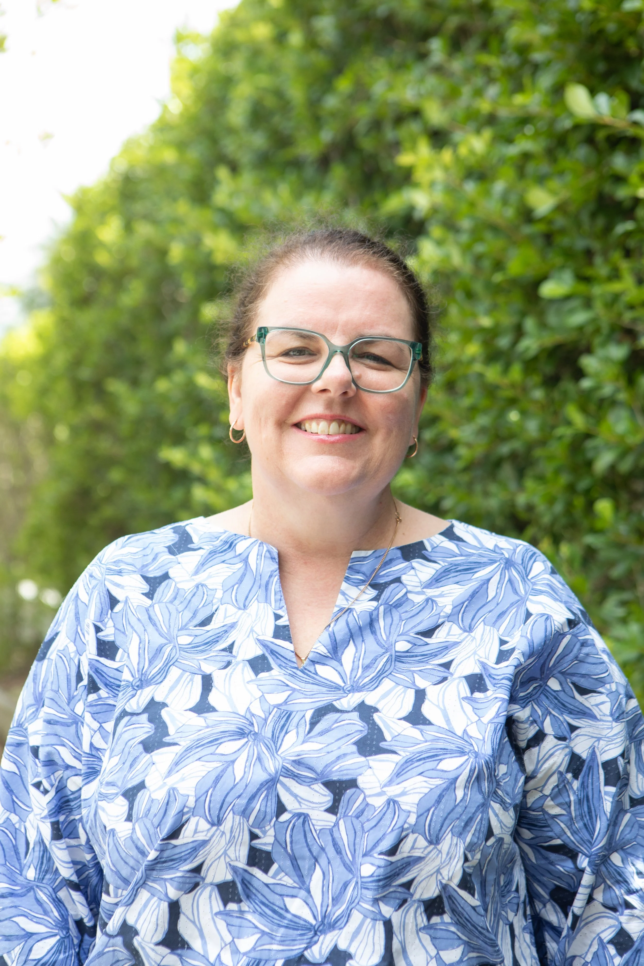 A smiling woman with glasses and earrings outdoors, wearing a blue and white floral top, with green foliage in the background.