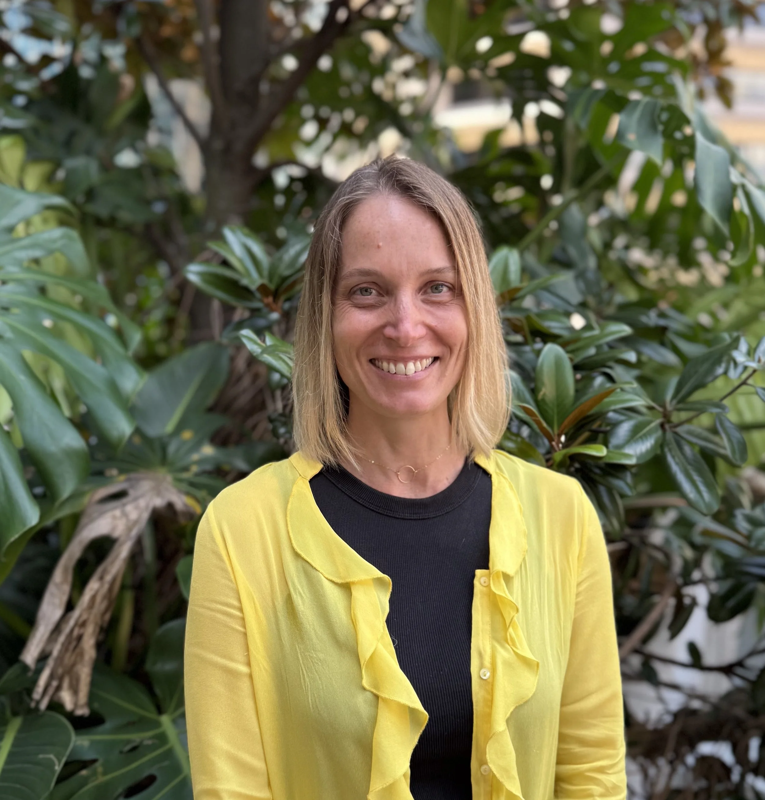 A woman with blonde hair smiling, wearing a yellow blouse and a black top, standing in front of green leafy plants.