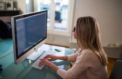 Woman working on a desktop computer with code on the screen in a well-lit room.