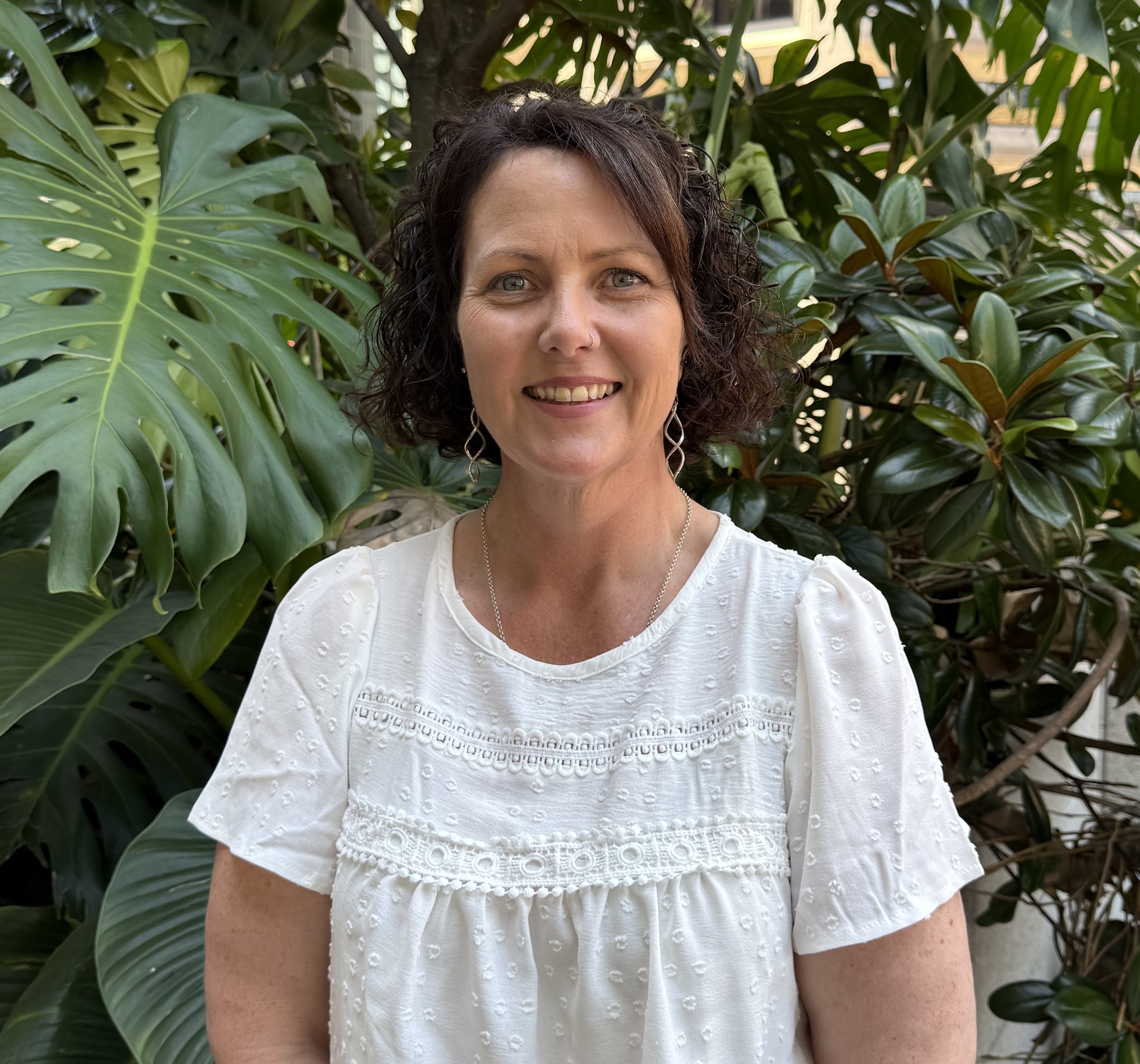 A woman with short, curly dark hair smiling, wearing a white embroidered blouse, standing in front of lush green tropical plants.