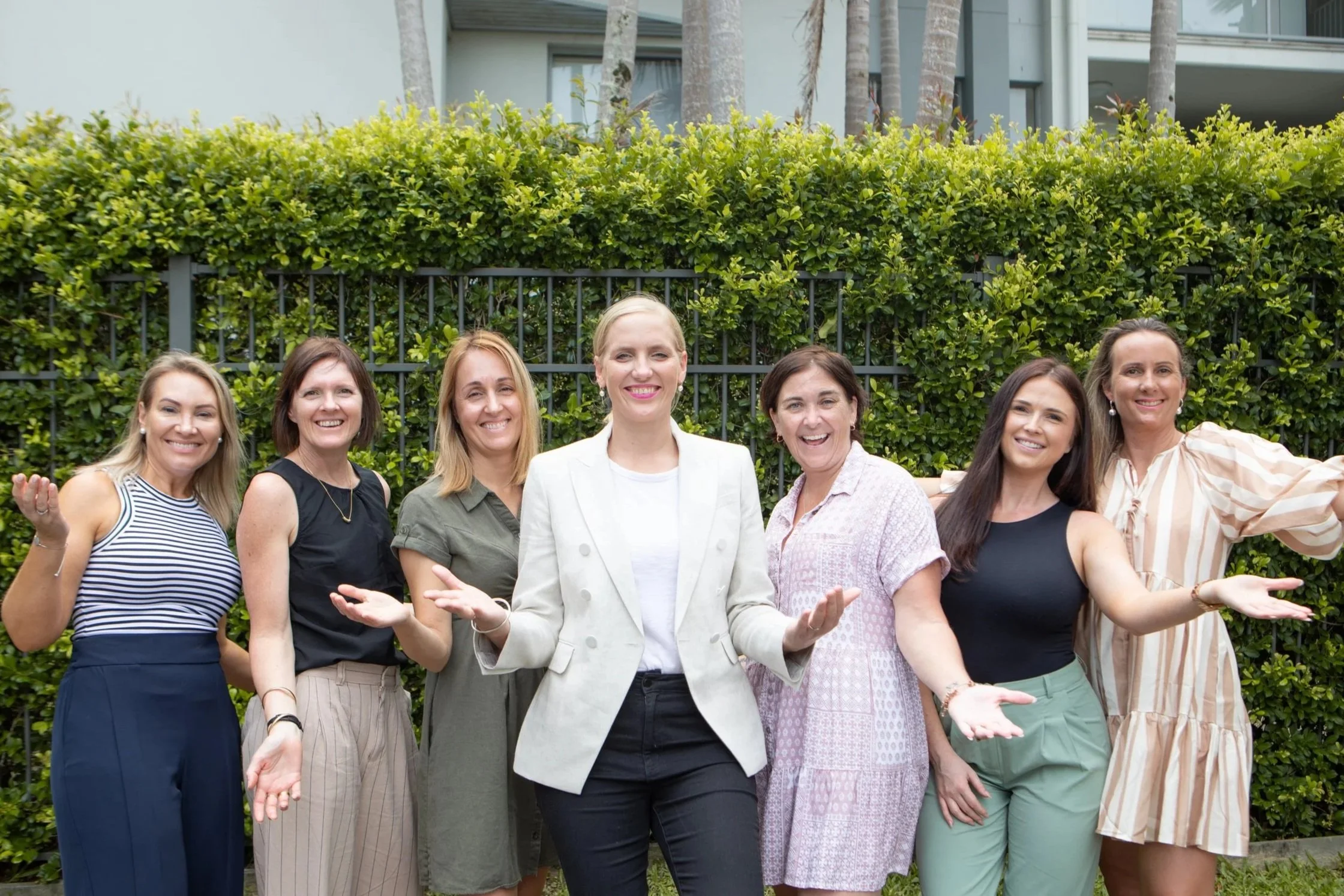 Group of seven women smiling and posing outdoors in front of a hedge.