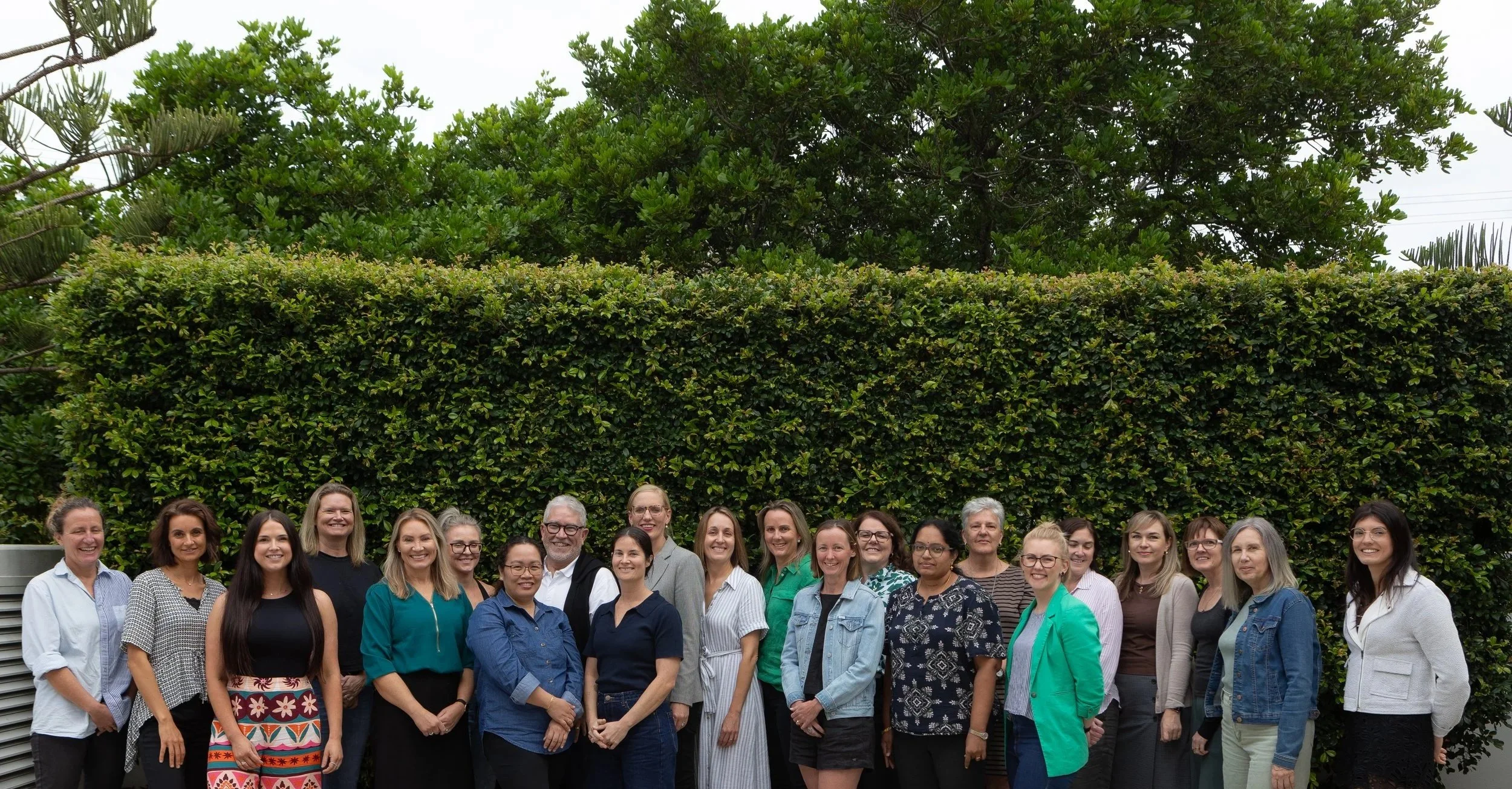 Group of diverse women with one man, smiling and standing outdoors in front of a green hedge