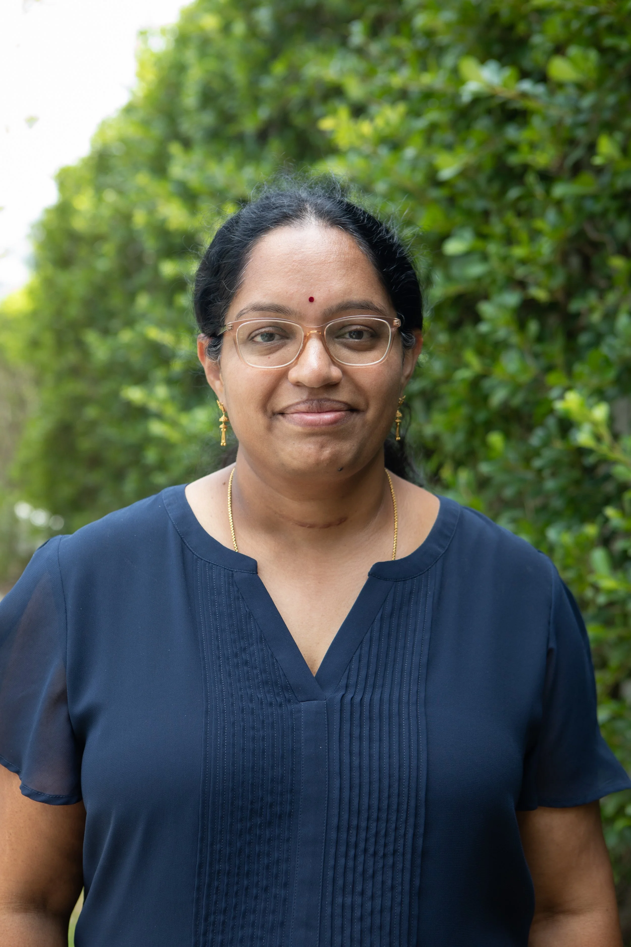 A woman of South Asian descent with glasses, gold jewelry, and a red bindi, standing outdoors with green foliage in the background.