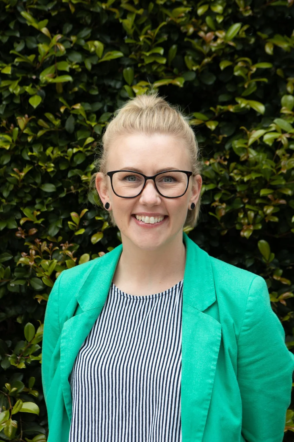 A woman with blonde hair tied back, wearing glasses, a turquoise blazer, and a striped shirt, smiling in front of a green leafy background.