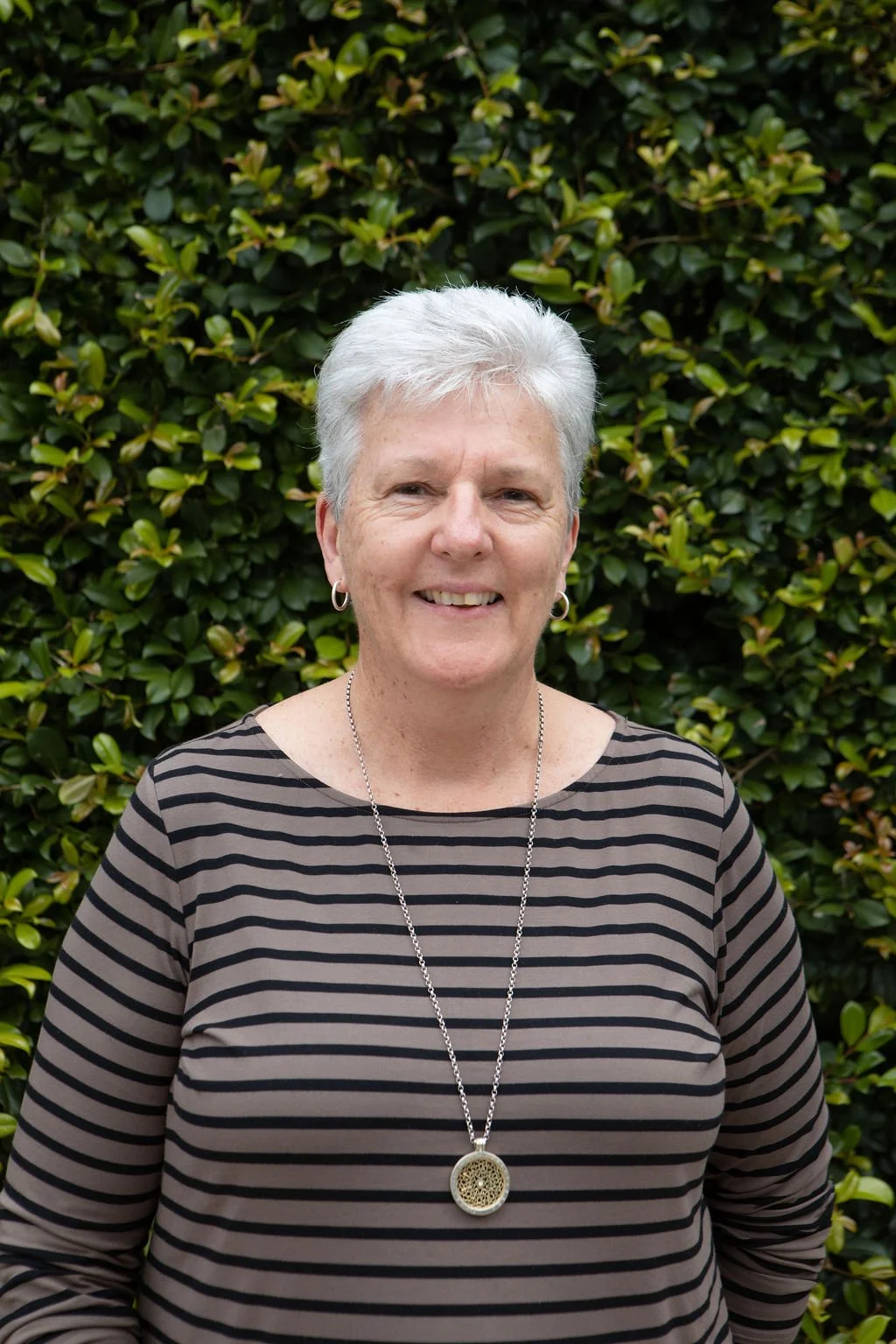 A smiling older woman with short gray hair standing outdoors in front of green leafy bushes, wearing a striped long-sleeved shirt and a pendant necklace.