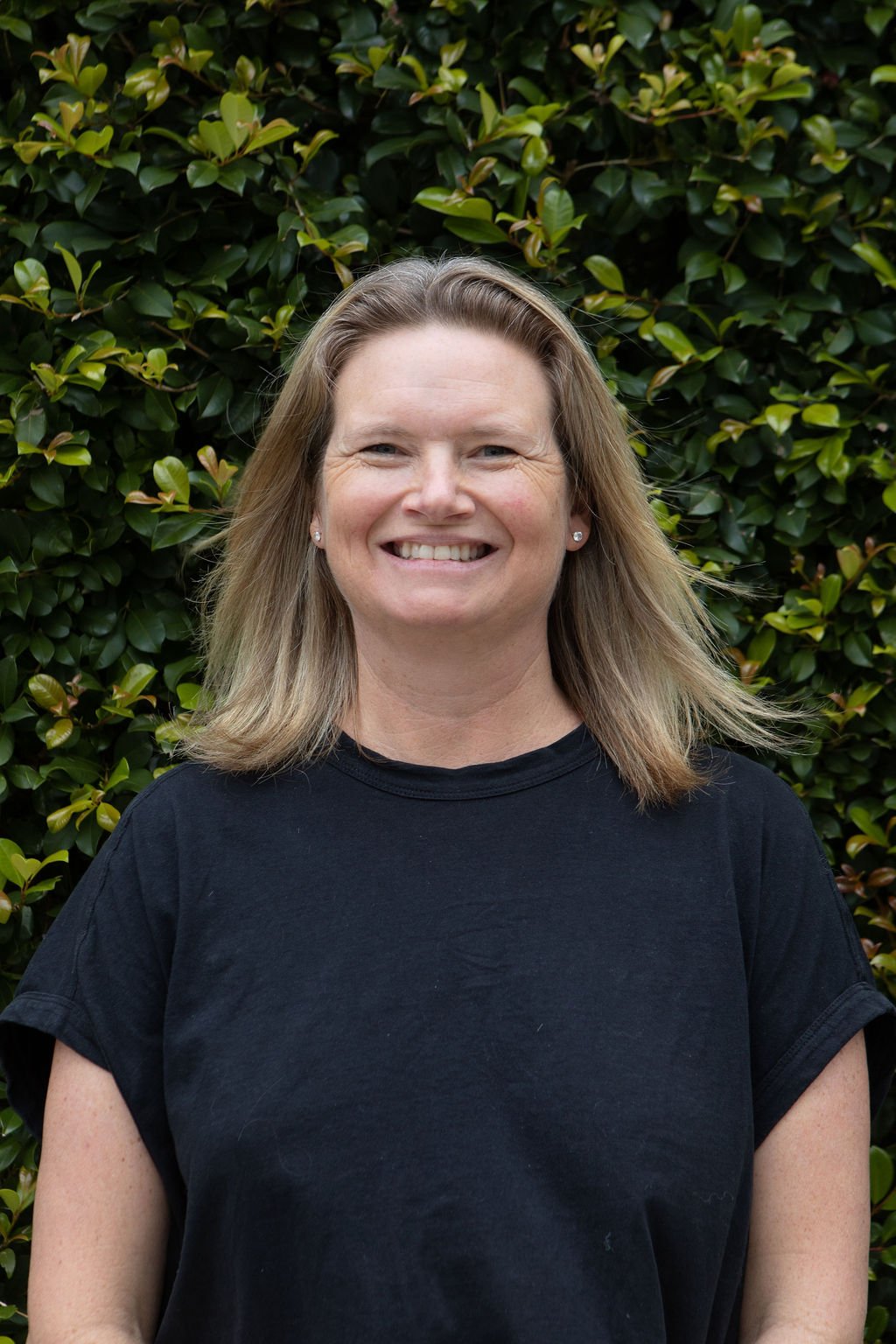 A woman with shoulder-length blonde hair, smiling, standing outdoors in front of green leafy bushes, wearing a black t-shirt.