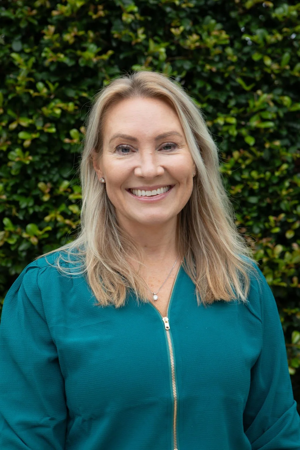 A woman smiling outdoors with green leafy background, wearing a teal zip-up jacket and a pearl necklace.