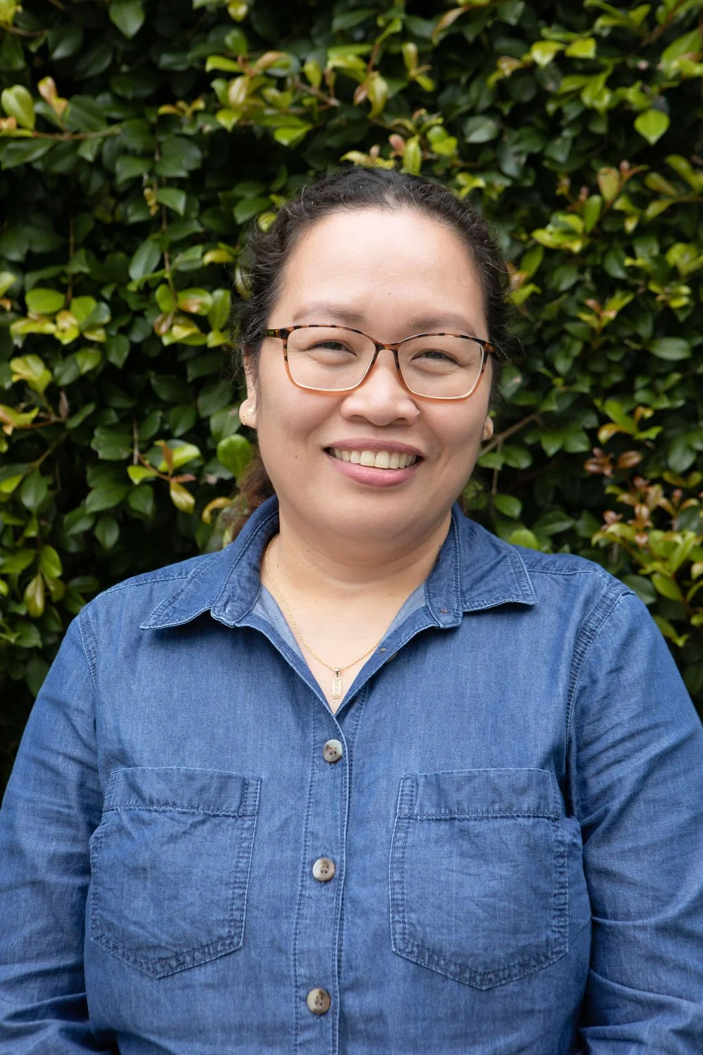 A woman wearing glasses and a blue denim shirt standing in front of a green leafy bush, smiling at the camera.