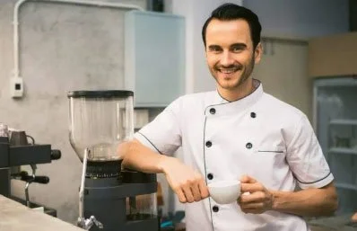 A male barista in a white chef's coat smiling while holding a white coffee cup in a coffee shop kitchen.