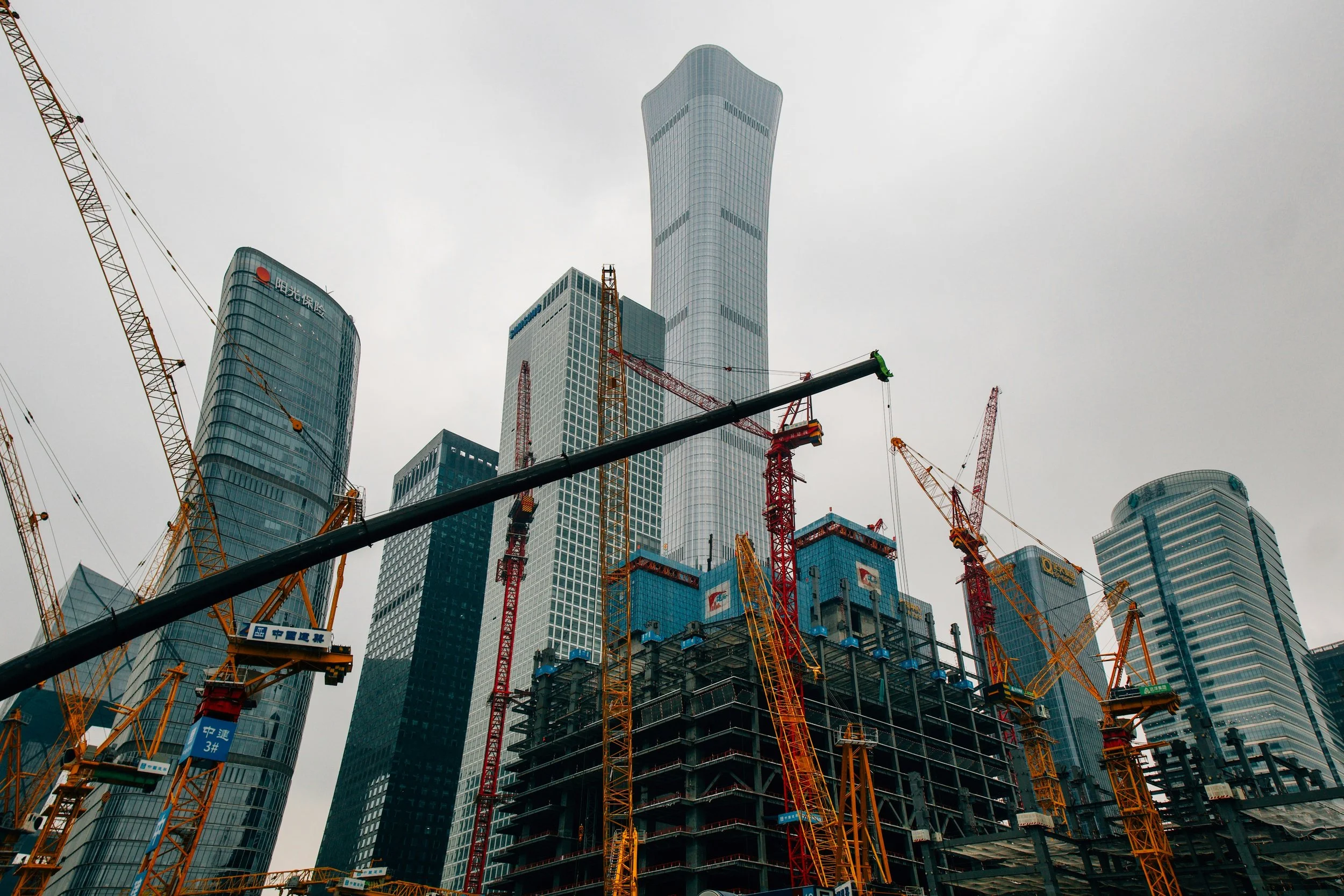 Construction site with multiple cranes and skyscrapers under cloudy sky.