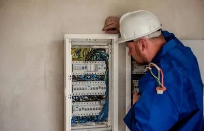 A male electrician working on an electrical panel in a white wall, wearing a white hard hat and blue uniform.