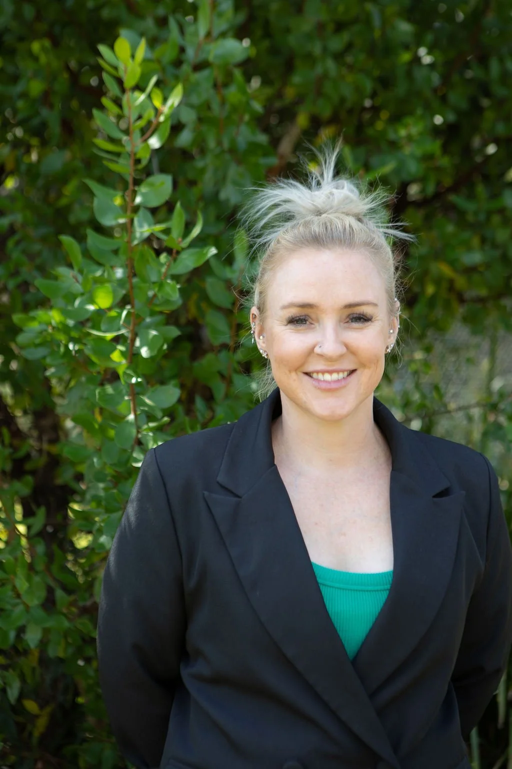 A woman with blonde hair tied in a messy bun, wearing a black blazer and a turquoise top, smiling outdoors in front of green foliage.