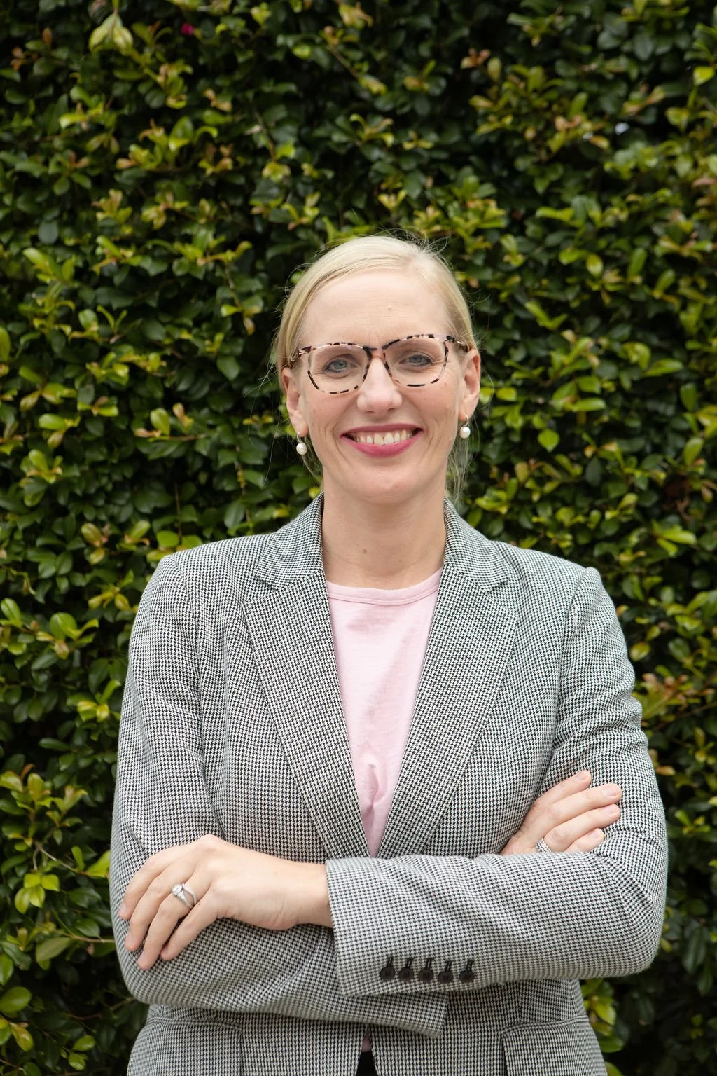 A smiling woman with blonde hair, wearing glasses, pearl earrings, a checkered blazer, and a pink shirt, standing with arms crossed in front of green leafy bushes.