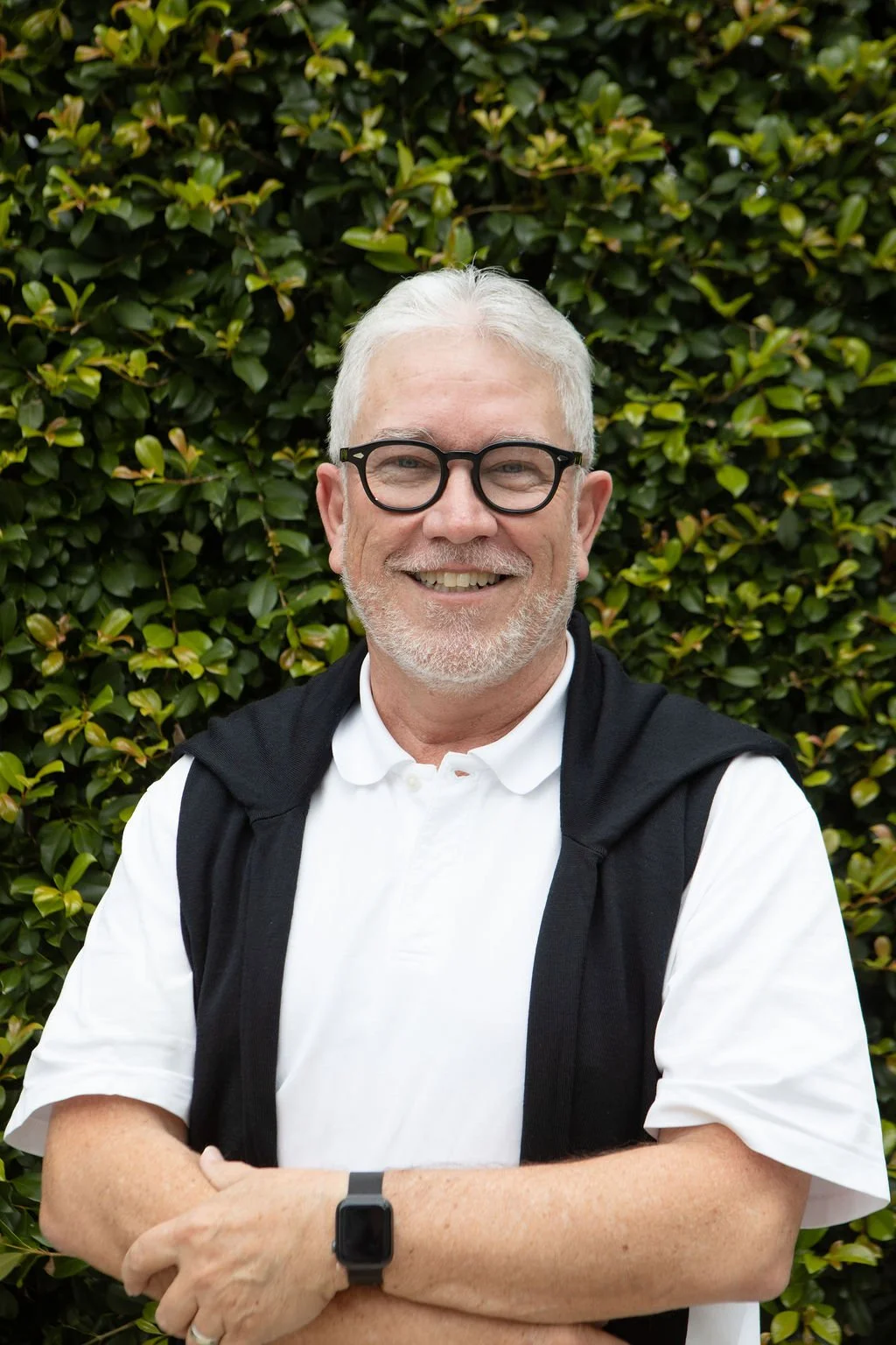 A smiling older man with gray hair and beard, wearing black-rimmed glasses, a white polo shirt, and a black jacket draped over his shoulders, standing in front of a leafy green hedge.