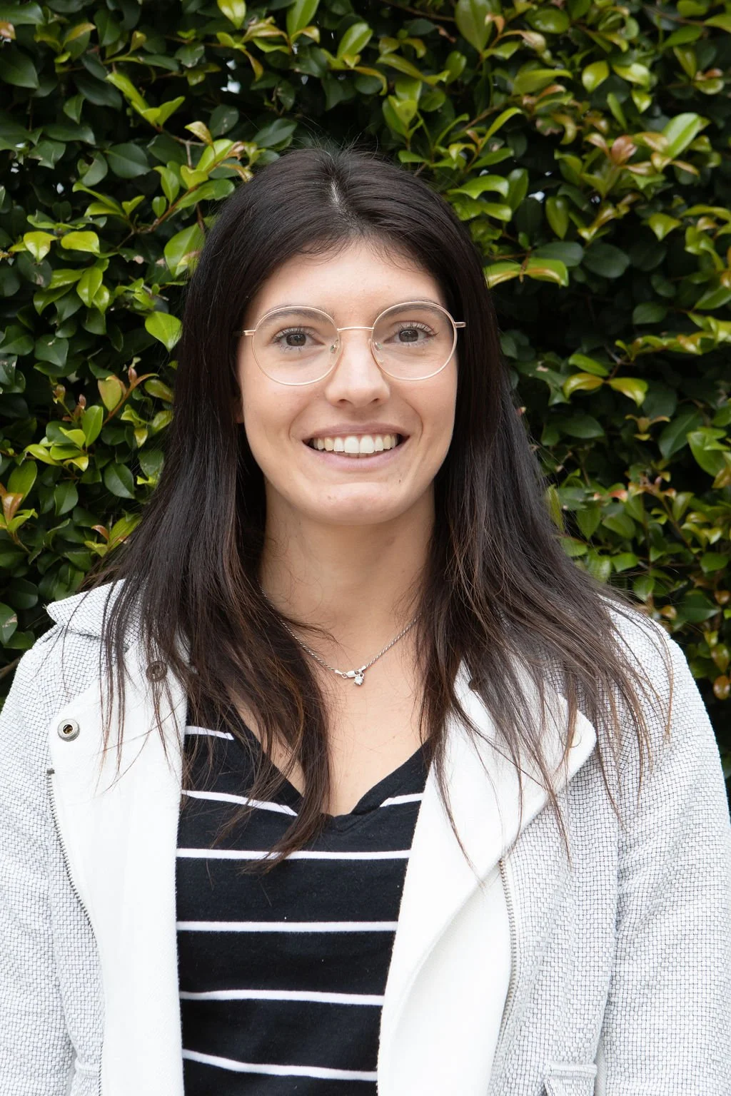 A young woman with long dark hair, glasses, and a striped black and white shirt, smiling outdoors in front of a green leafy bush.