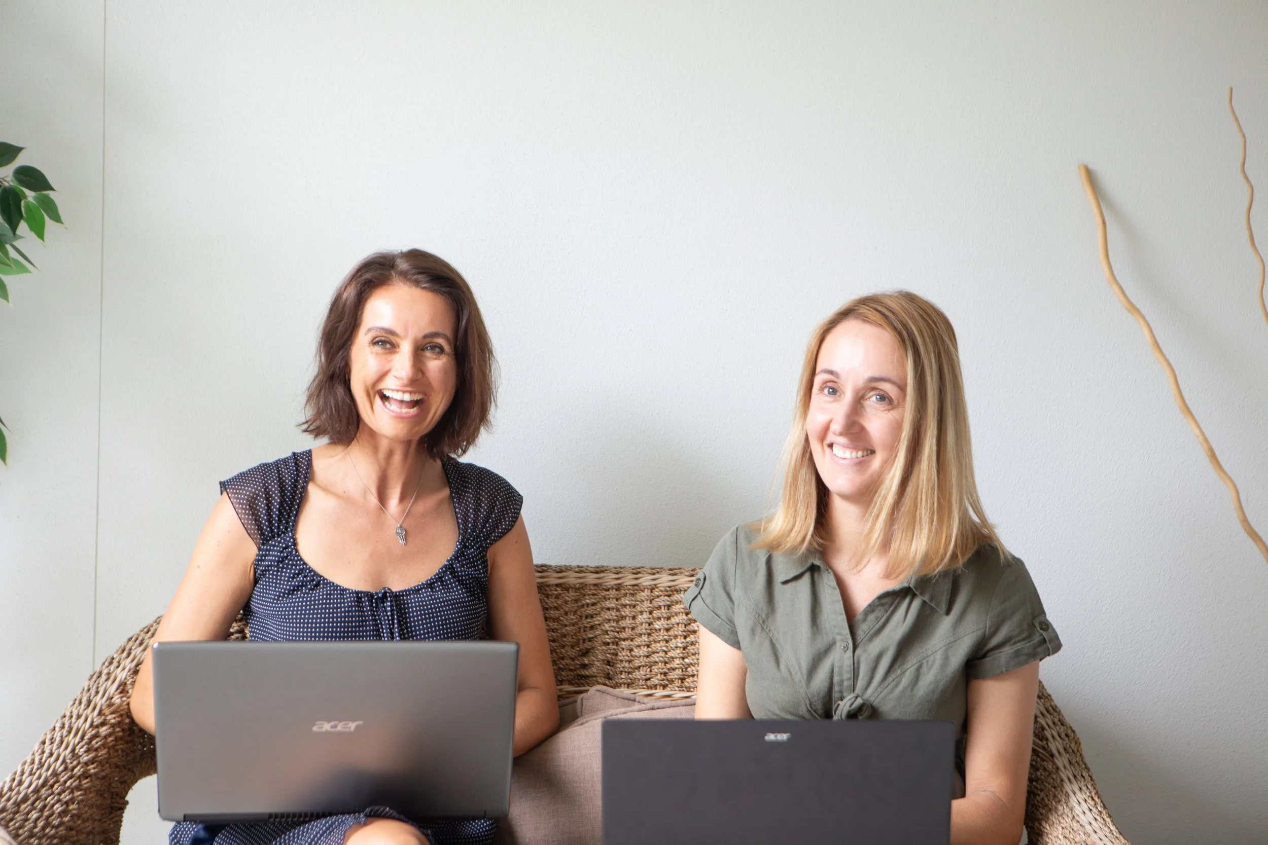 Two women sitting on a wicker couch, each with a laptop, smiling and looking happy, in a minimalistic room with a white wall, a green plant on the left, and decorative branches on the right.