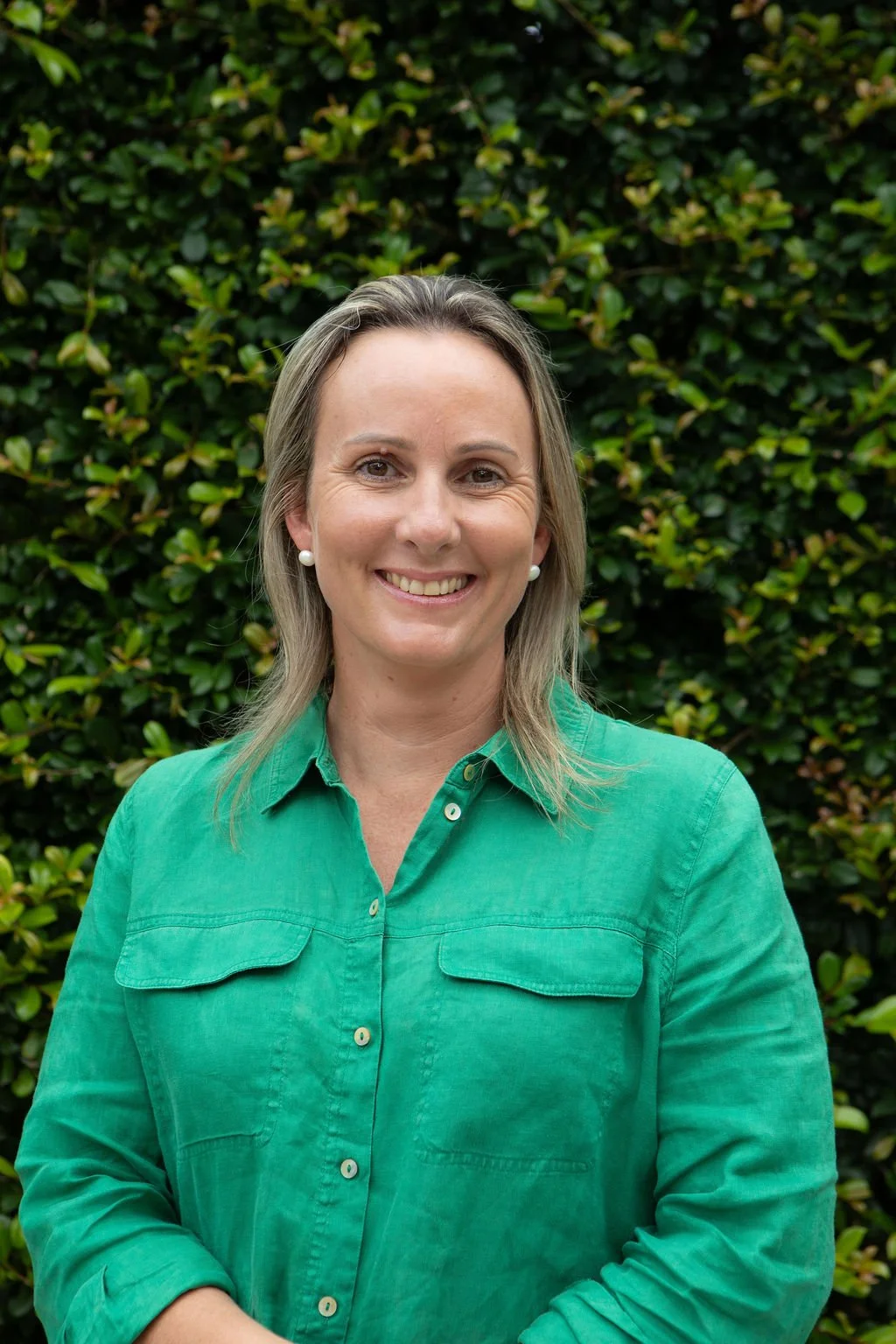 A woman with shoulder-length blonde hair wearing a green button-up shirt smiling in front of a leafy green background.