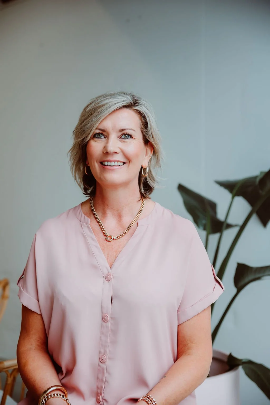 A woman with short blonde hair, wearing a pink blouse, gold jewelry, and smiling, standing indoors near a large potted plant.