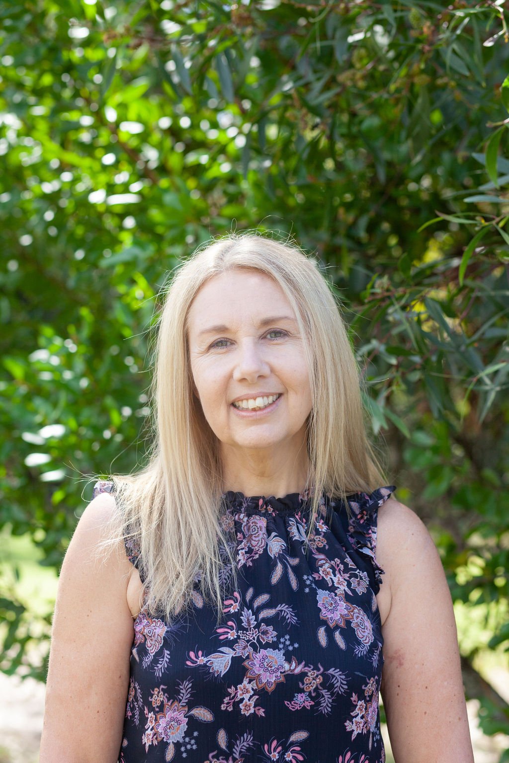 A woman with long blonde hair smiling outdoors, standing in front of green leafy foliage.