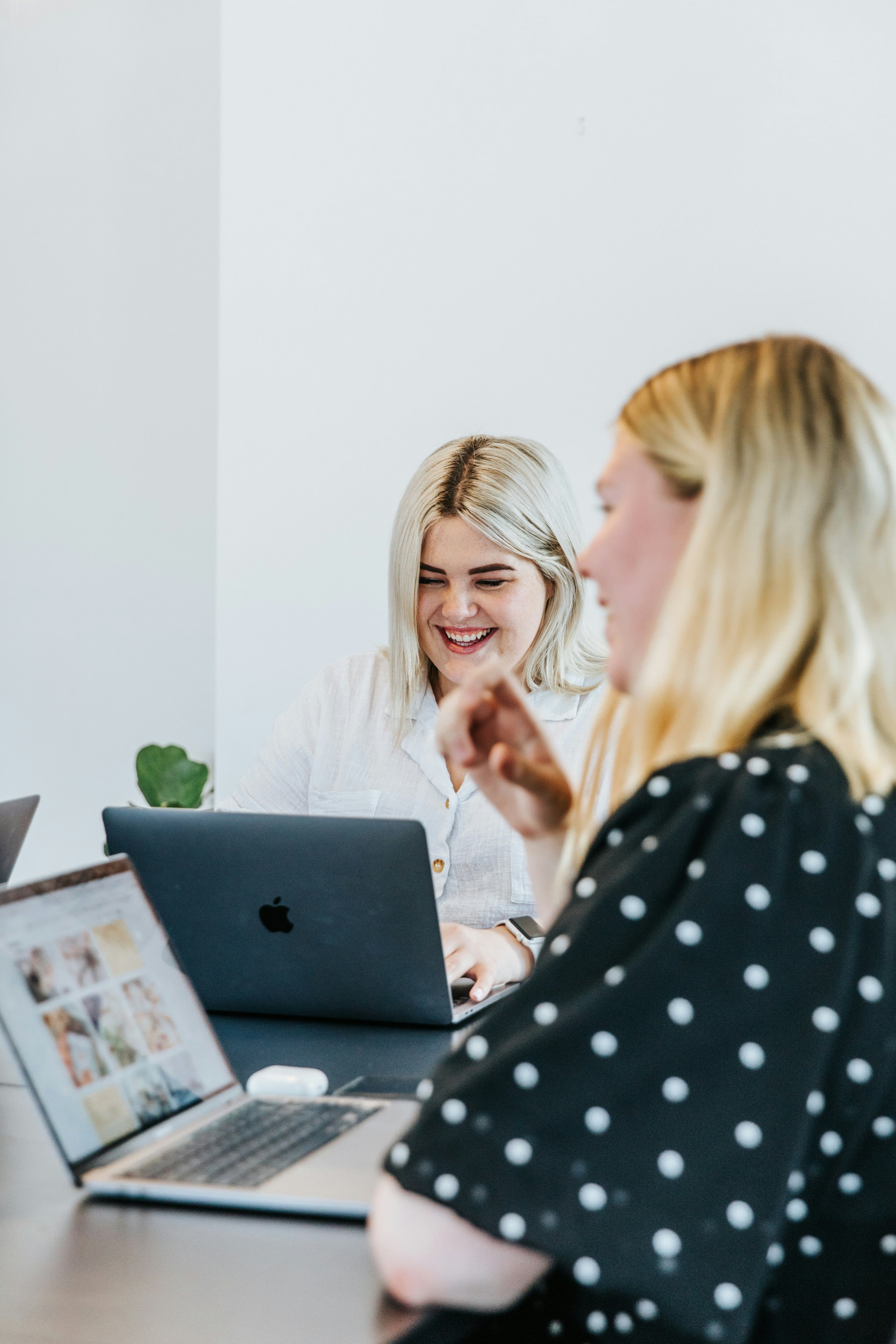 Two women are seated at a table working on laptops in a bright, minimalistic office space, one smiling and looking down at her device.