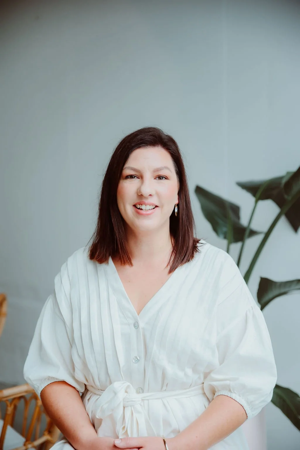 Smiling woman with shoulder-length brown hair and earrings, wearing a white dress, sitting indoors near a large green plant.