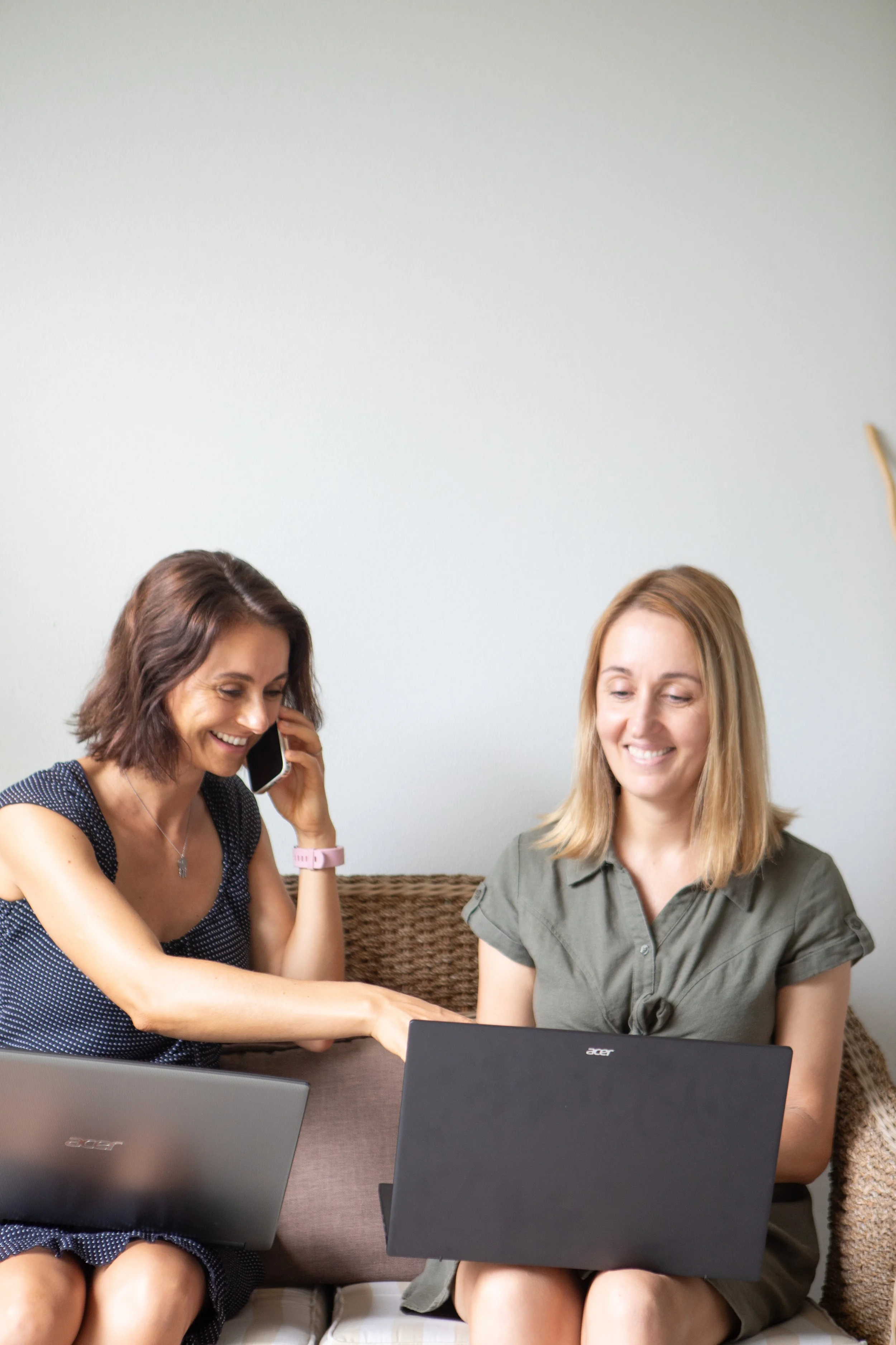 Two women sitting on a sofa, working on laptops and talking on the phone, in a casual indoor setting.