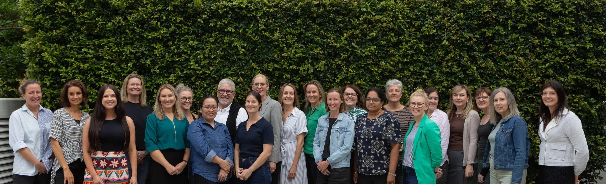 Group of diverse people standing together outdoors in front of green hedge, smiling for a photo.